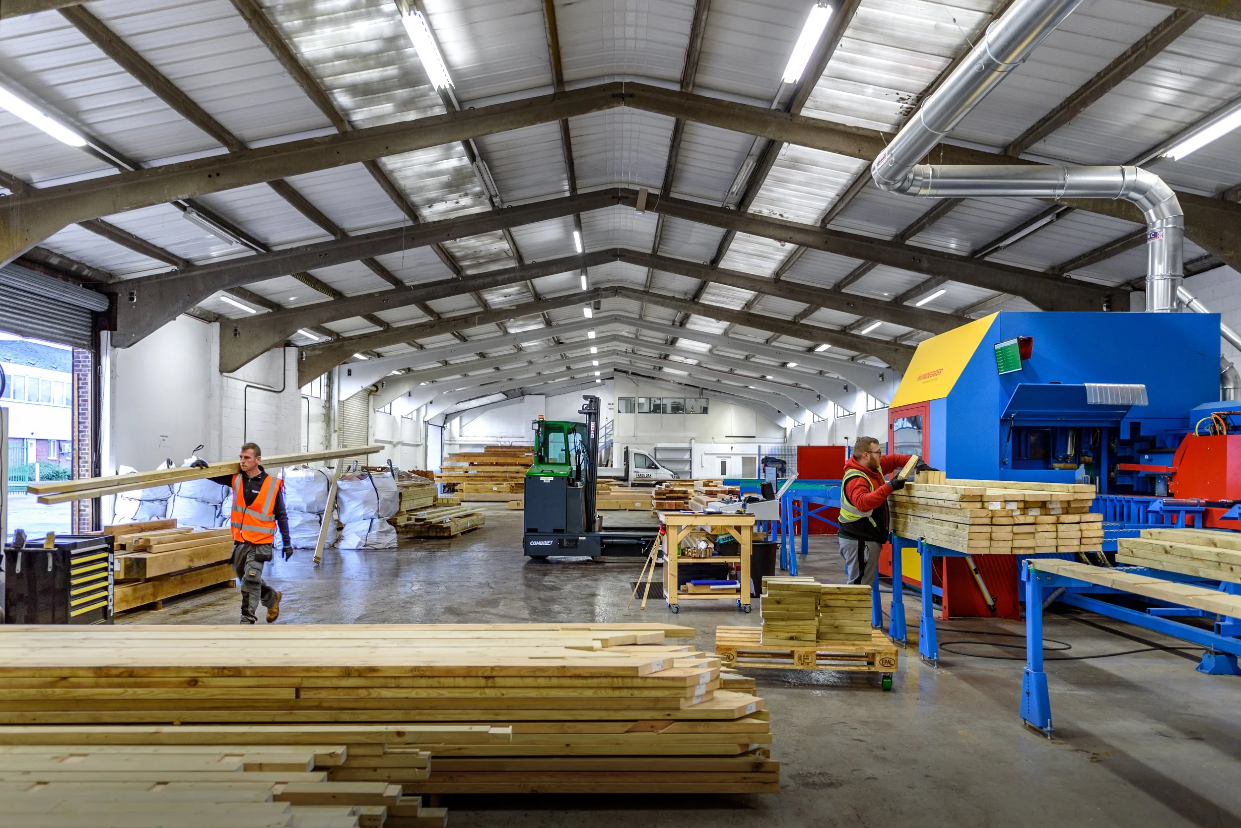 Inside a woodworking factory with workers handling lumber, a large machine, and stacks of wood on pallets and tables.