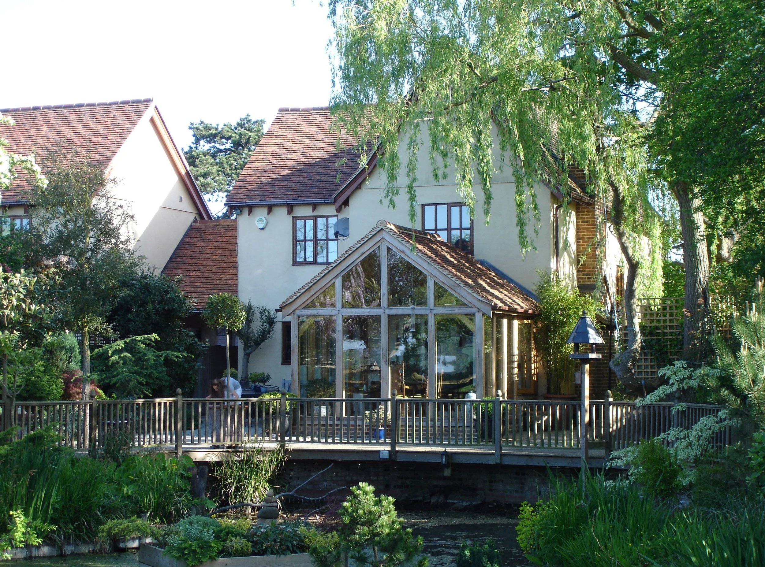A house with white walls and a brown tiled roof, surrounded by lush green trees and bushes. There is a wooden deck with a glass greenhouse structure attached to the house, and a small pond with plants in front.