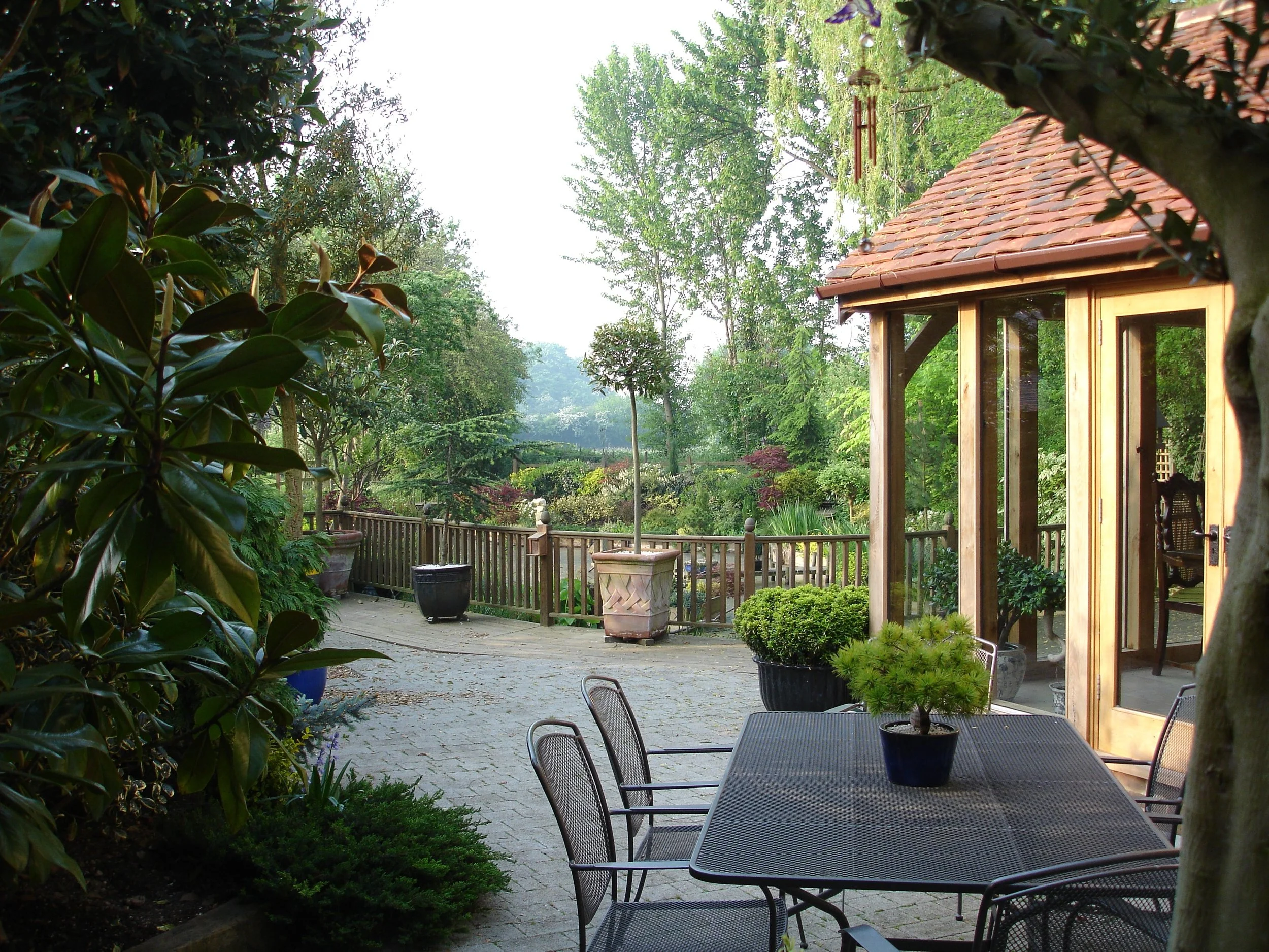 A patio garden with a black metal table and chairs, potted plants, and a wooden enclosed porch surrounded by lush green trees and plants.