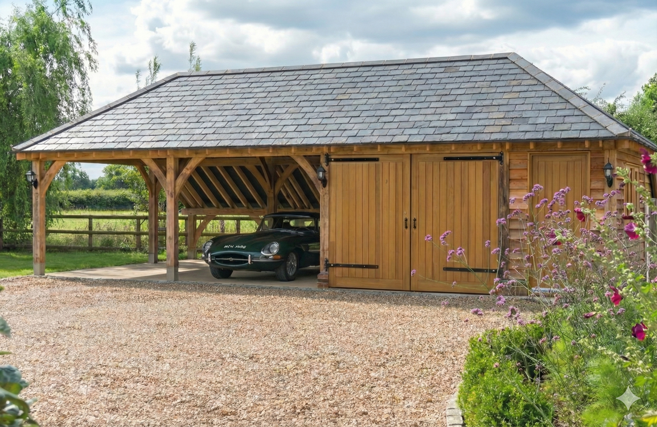 A wooden garage with sliding doors, houses a vintage dark green Jaguar car. The garage is situated in a lush, green outdoor setting with a gravel driveway and flowering plants nearby.