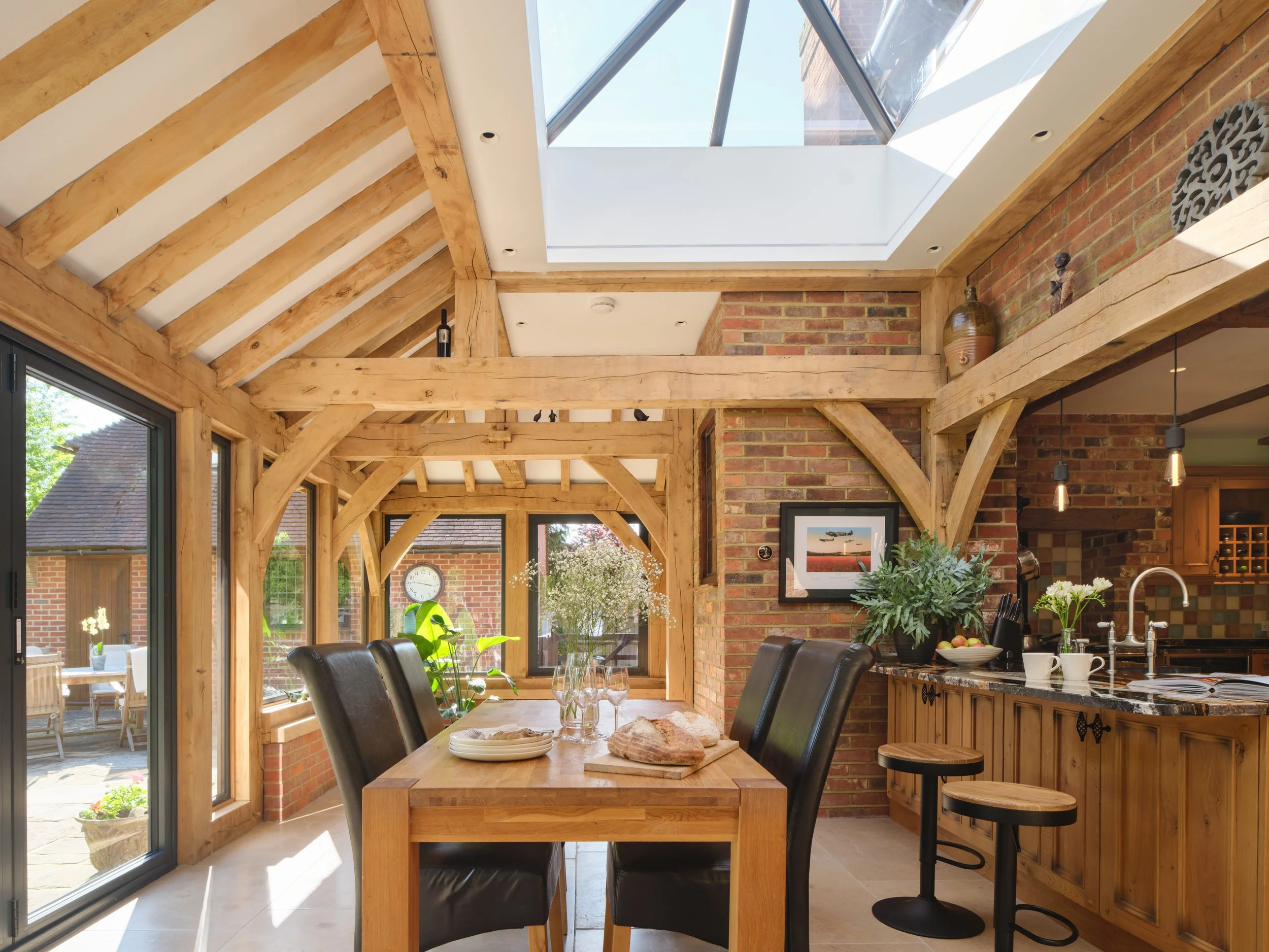 Interior view of a dining area with a wooden table, black chairs, and a kitchen counter with hanging lights. Large windows and a glass ceiling let in natural light. The space features exposed wooden beams and brick walls, decorated with plants and framed pictures.
