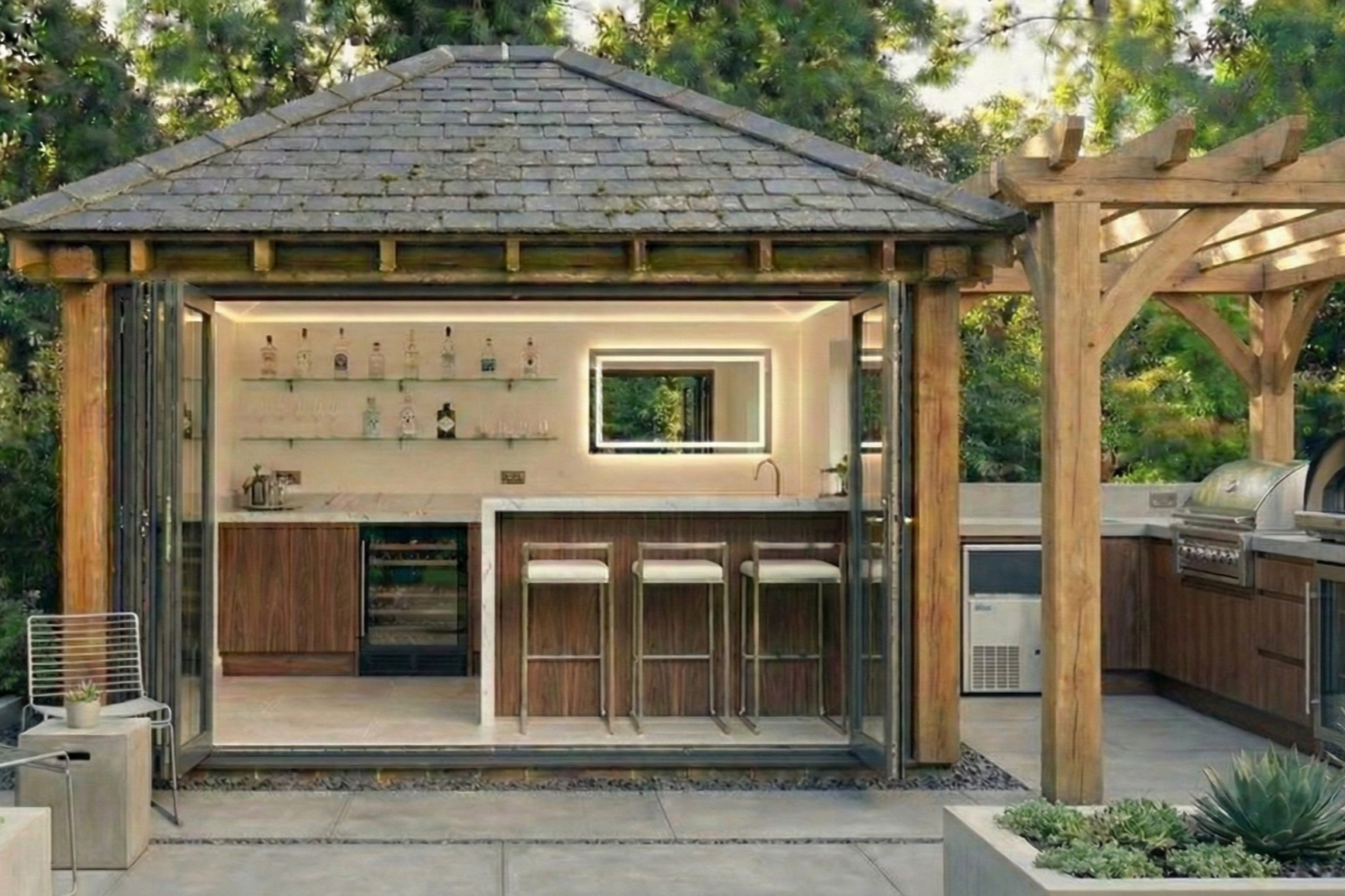 Outdoor bar area with a countertop, bar stools, and shelves of liquor bottles, enclosed by a glass structure with a shingle roof, surrounded by greenery.