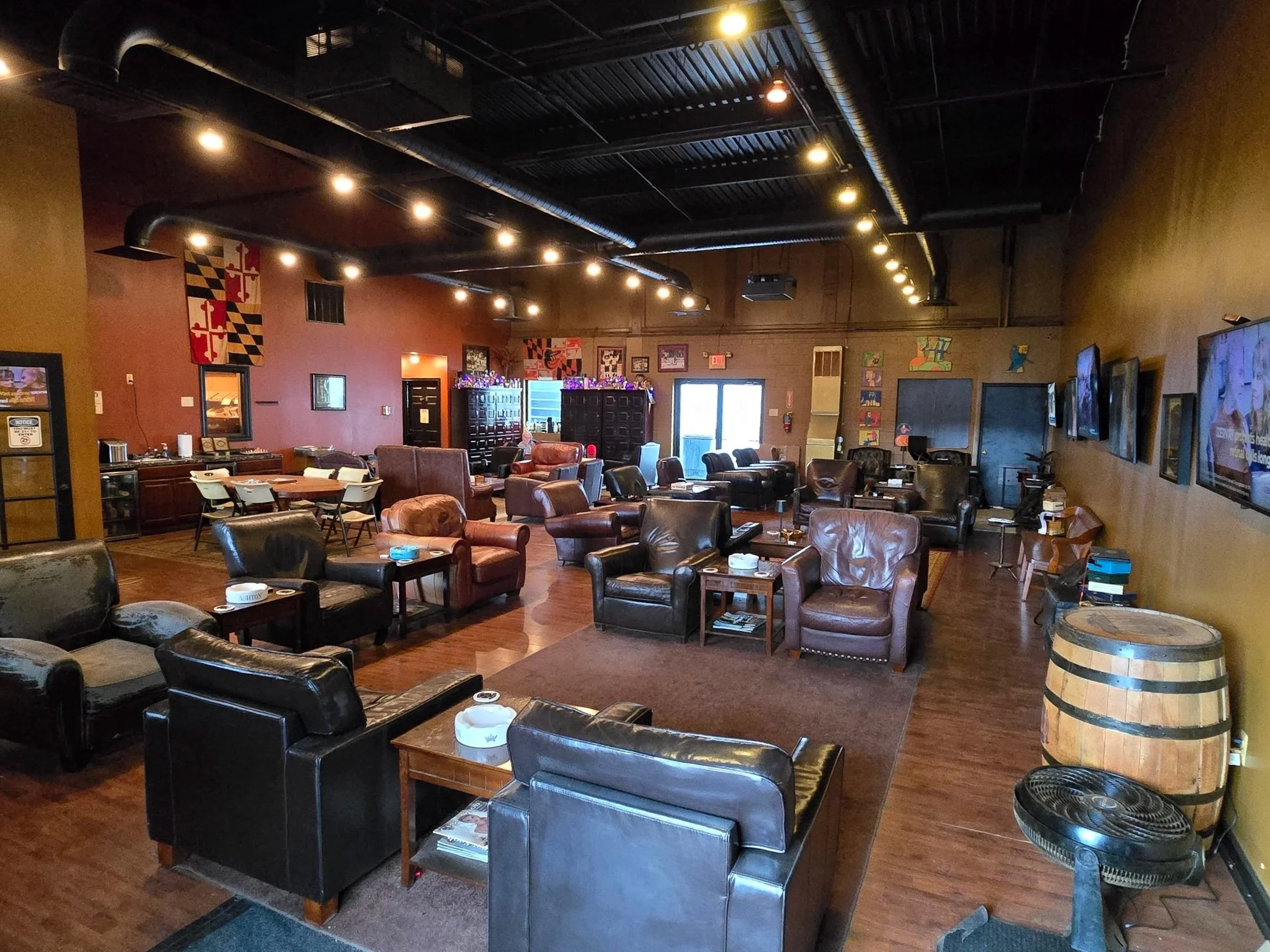 A spacious lounge area with leather chairs and sofas, wooden coffee tables, and TV screens on the walls, decorated with Maryland flags and artwork, with hardwood floors and warm lighting.