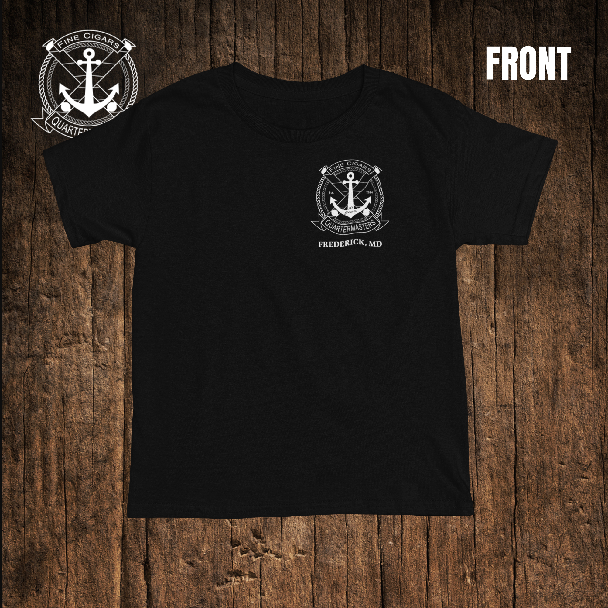 Black T-shirt with a white logo on the front and a logo on the left sleeve. The front logo features an anchor with a rope, surrounded by a circular rope border with banners and the words "Fine Cigars" and "Quartermasters." Below the logo on the front, it reads "Frederick, MD." The background is a wooden surface.
