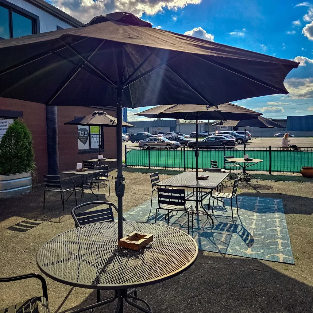 An outdoor patio with metal tables and chairs, large black umbrellas, and a colorful rug, with a parking lot and blue sky with clouds in the background.