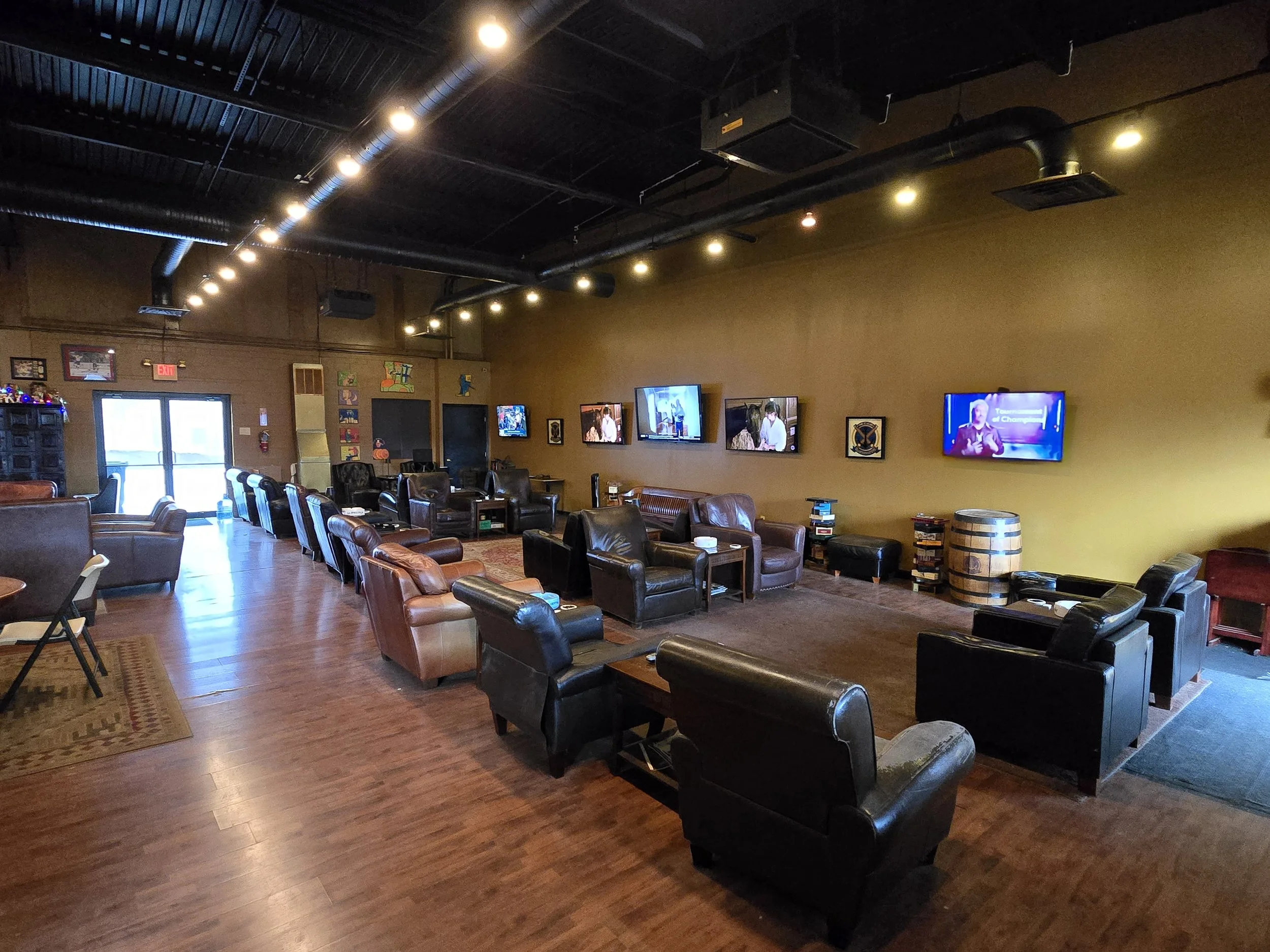 A cozy lounge with multiple leather chairs and sofas arranged in clusters, several flat-screen televisions on a mustard-colored wall, and a wooden barrel used as a table, with hardwood flooring and exposed black ceiling pipes.