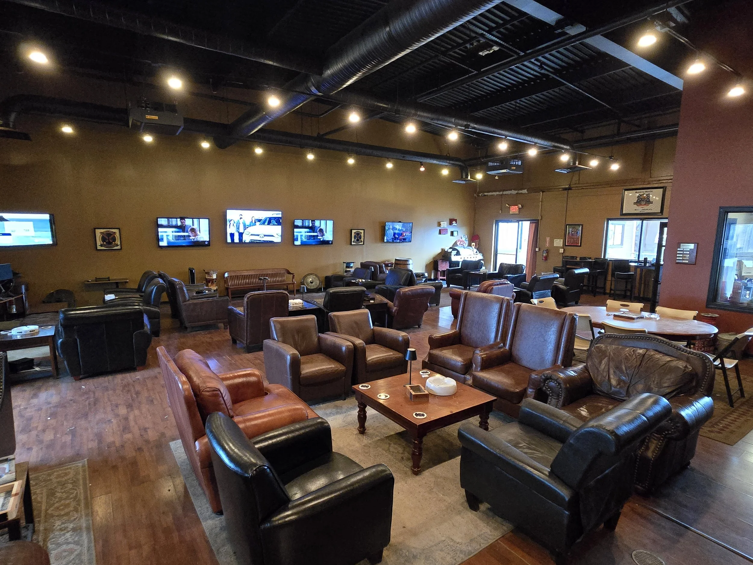 A cozy lounge area with leather chairs and couches, multiple wall-mounted TV screens, and hardwood floors, creating a comfortable atmosphere suitable for relaxation or socializing.