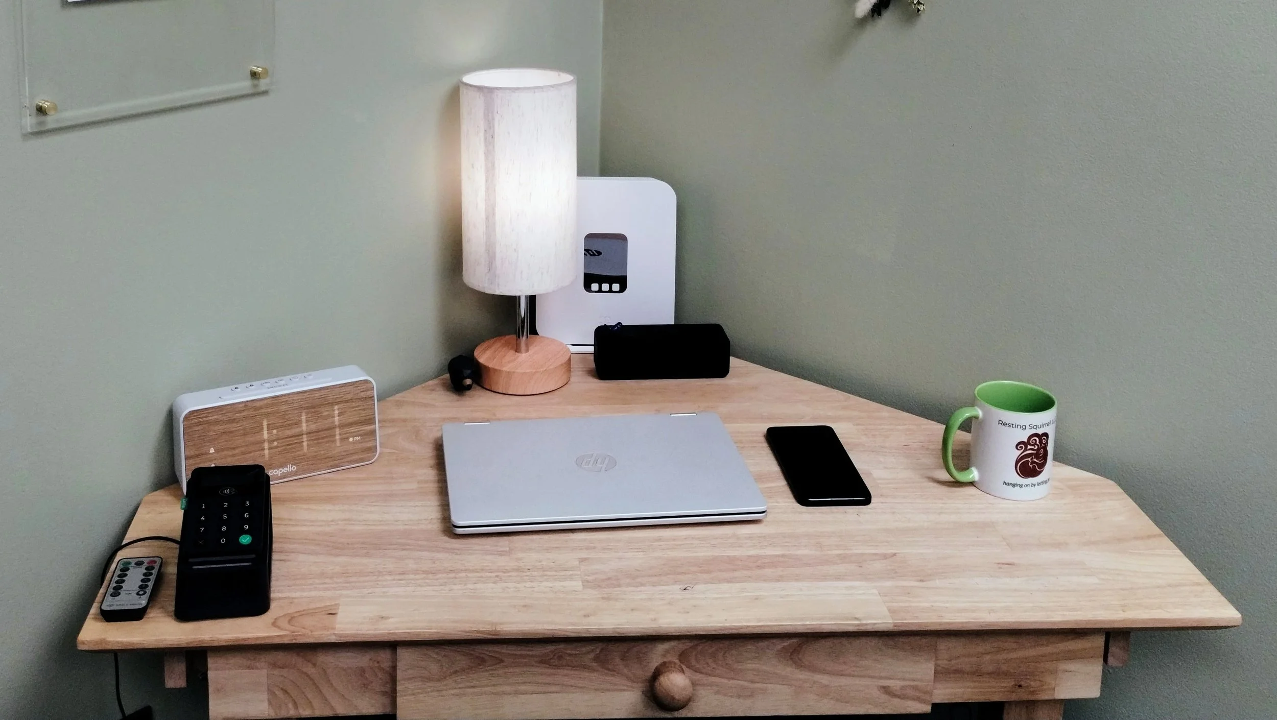 A very neat wooden corner desk at Resting Squirrel in Merrimack NH. It has a laptop, cellphone, speaker, lamp and modem.  And next to the lamp a small wooden bear looks on.