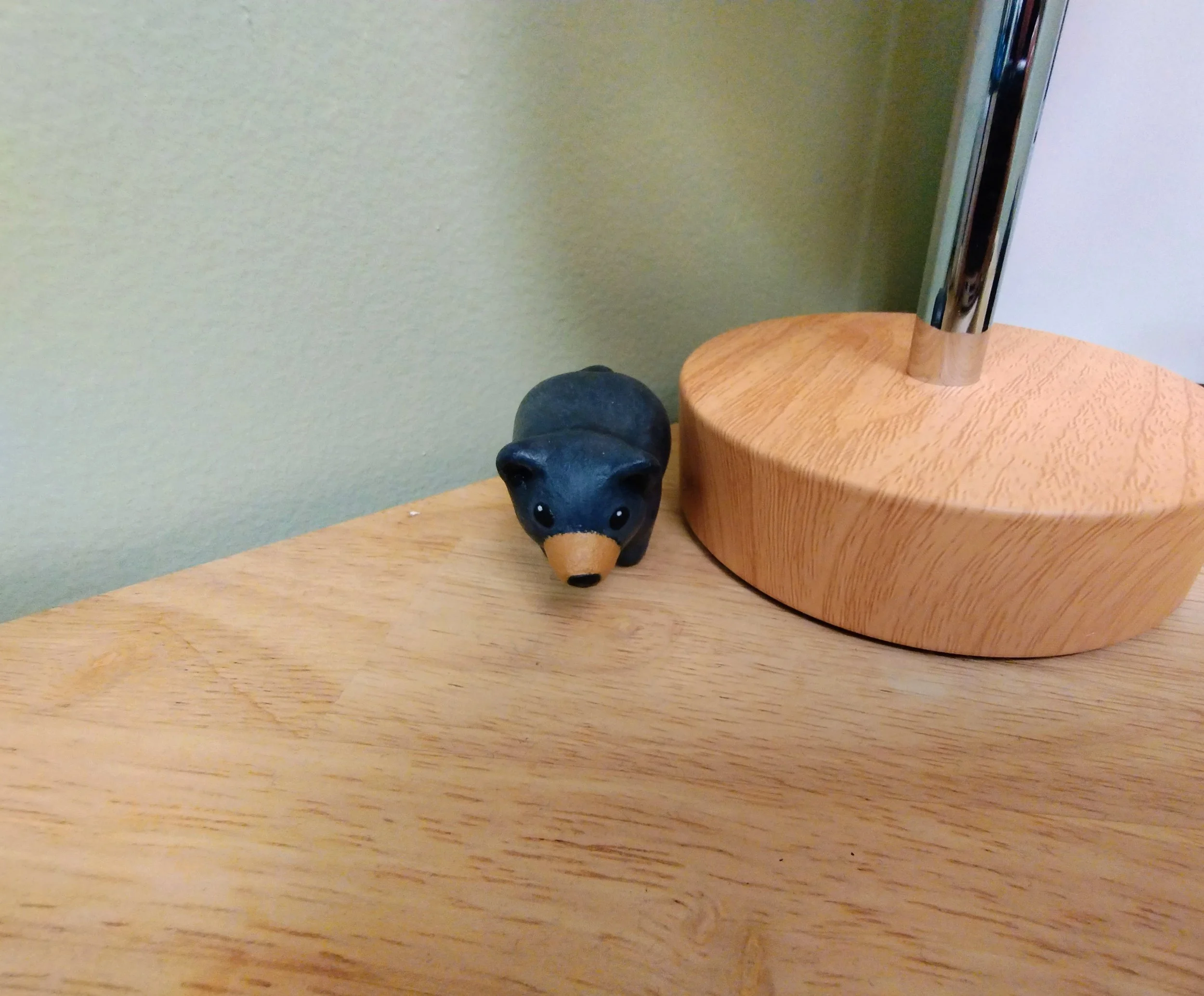 A small decorative black bear figurine with a tan nose, placed on a wooden surface next to a lamp with a wooden base and metal pole.