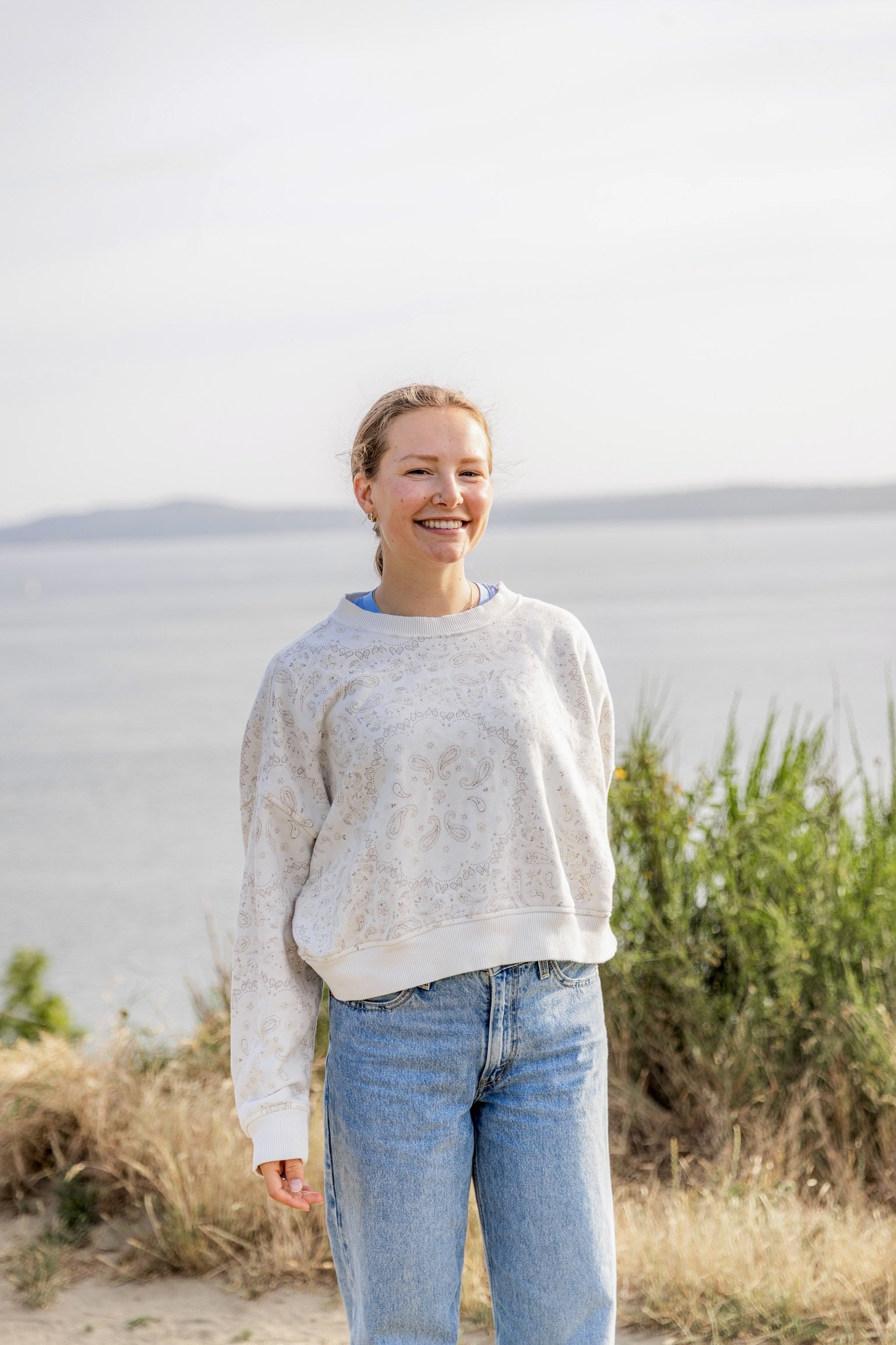 A young woman with blonde hair and a nose ring smiling outdoors near a body of water and greenery, wearing a white patterned sweatshirt and blue jeans.
