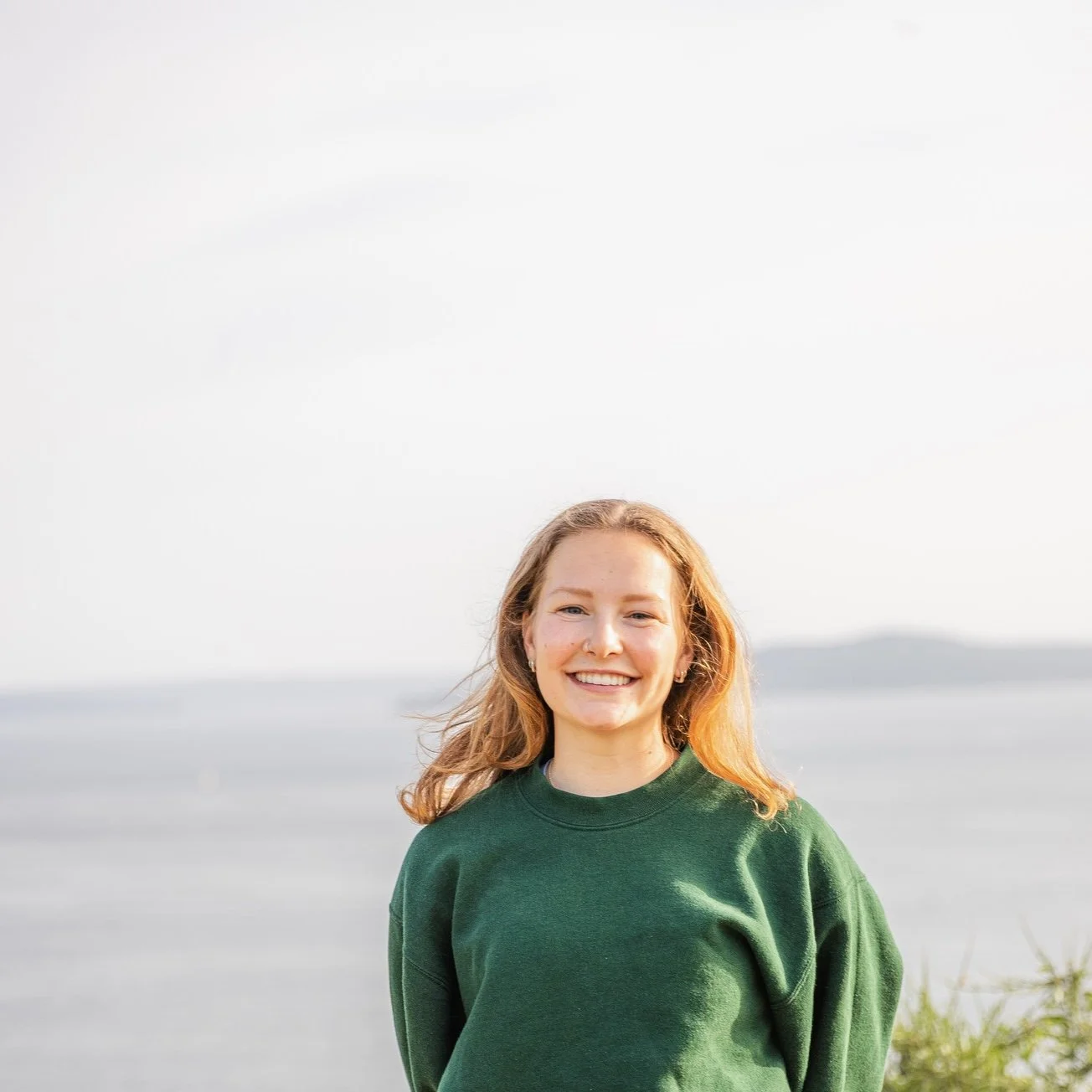 A young woman with red hair smiling outdoors near a body of water, wearing a green sweatshirt.