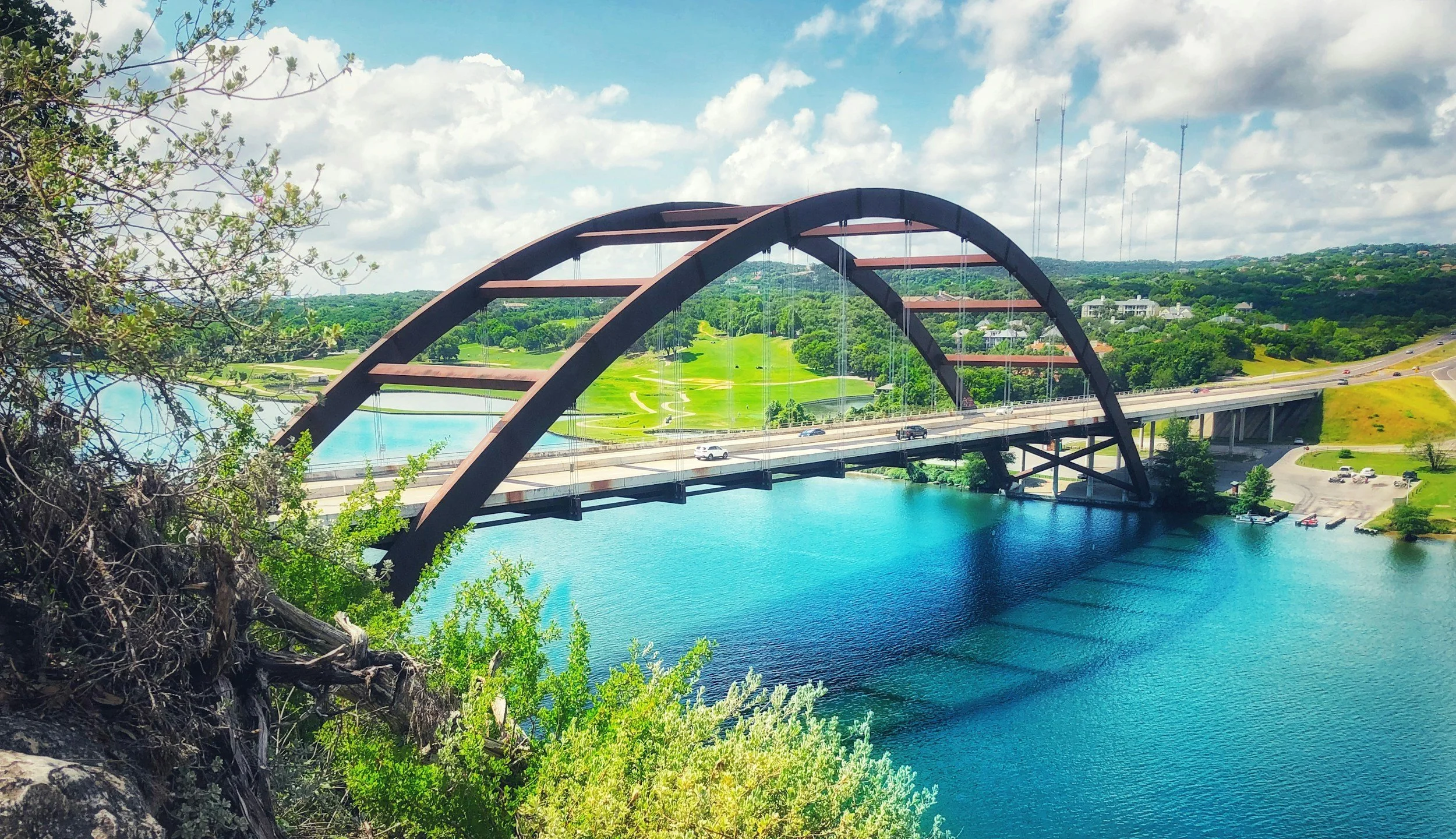 A scenic view of a bridge with large arch supports over a body of water, with green hills and blue sky in the background.