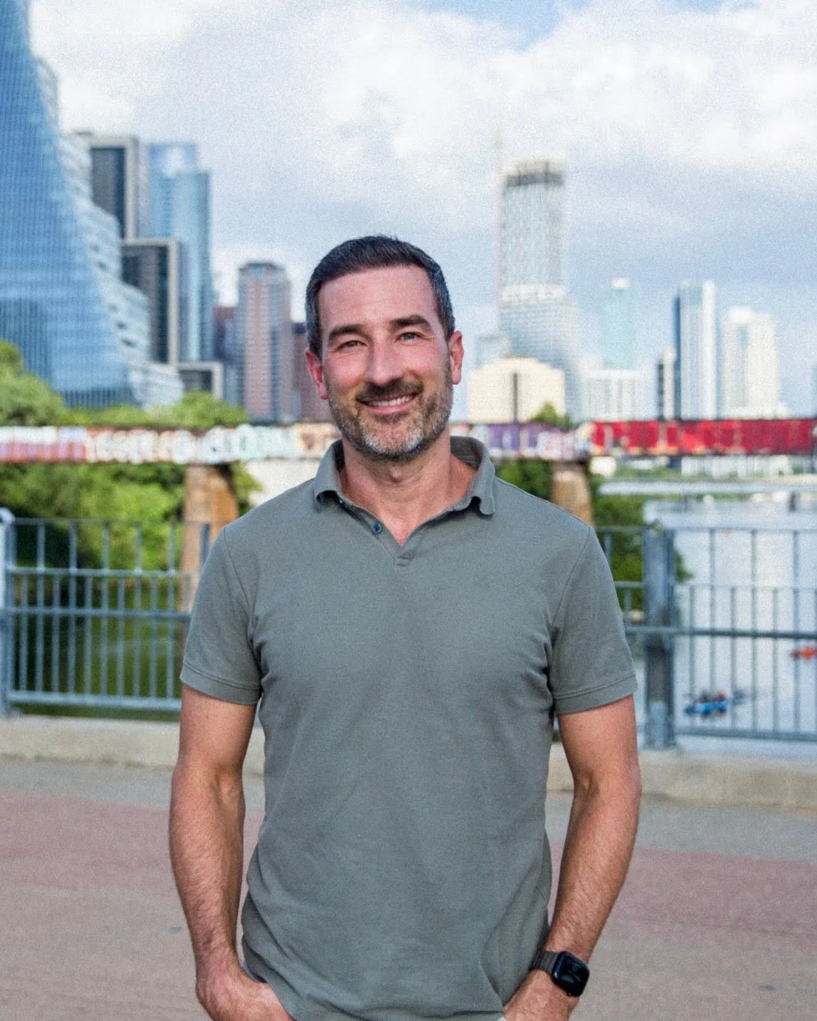 A smiling man with dark hair and a beard stands outdoors in front of a city skyline with tall modern buildings and a river, wearing a gray polo shirt and a smartwatch.