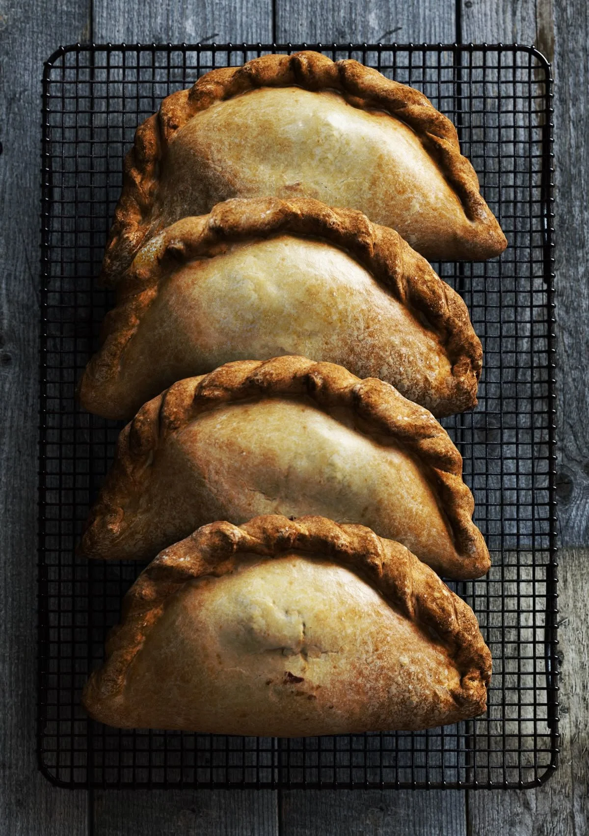 Four baked turnovers with golden-brown crusts arranged in a row on a wire cooling rack, against a rustic wooden background.