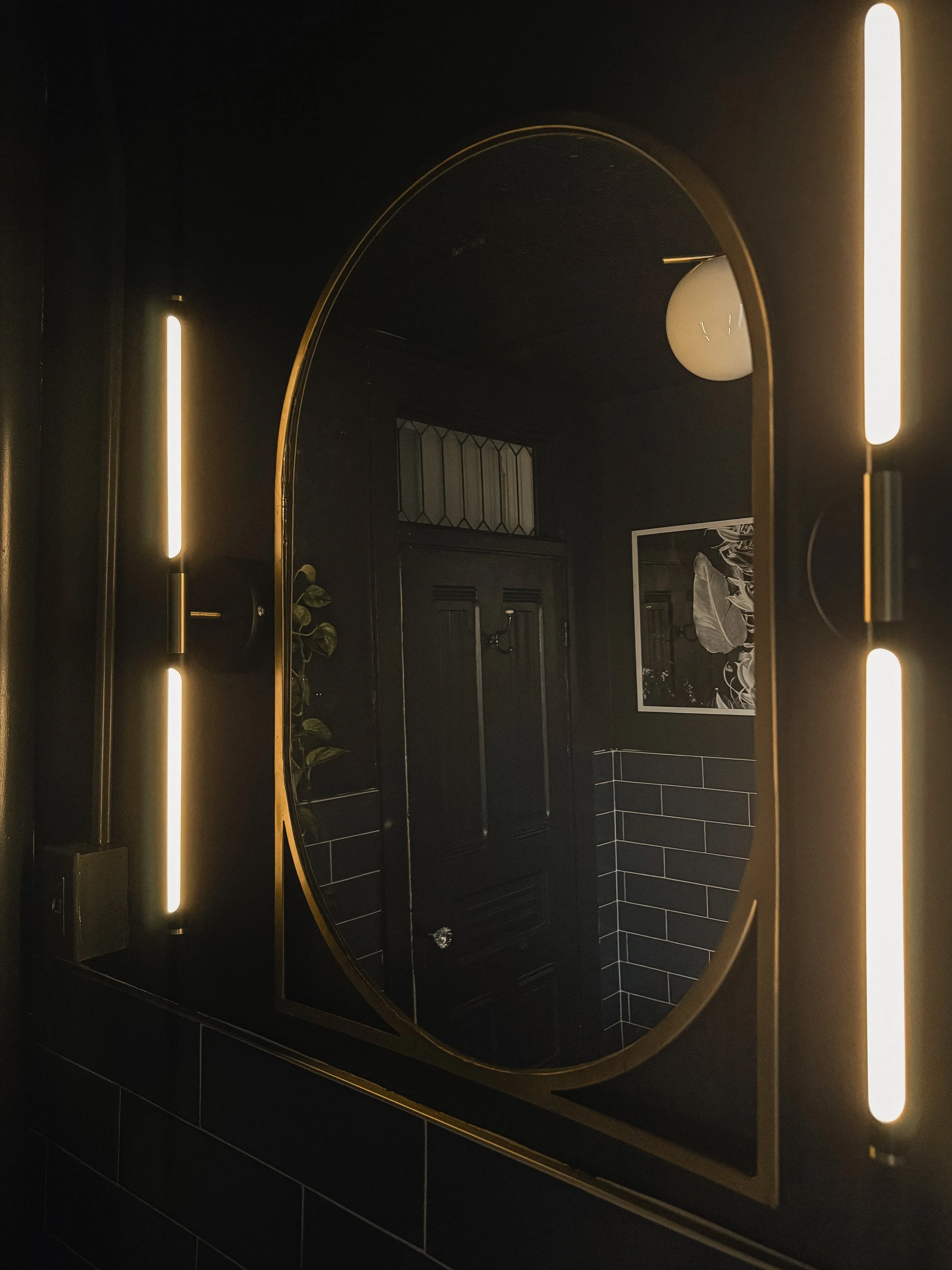 Moody bathroom at the West Temple residence with an illuminated oval mirror, dark walls, and vertical wall lighting.