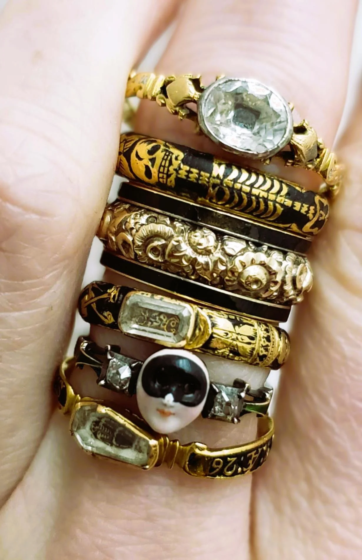 Close-up of a finger with multiple gold rings from the late 17th century to the 18th century, including a rare masquerade souvenir ring and a rare skeleton-in-coffin ring.