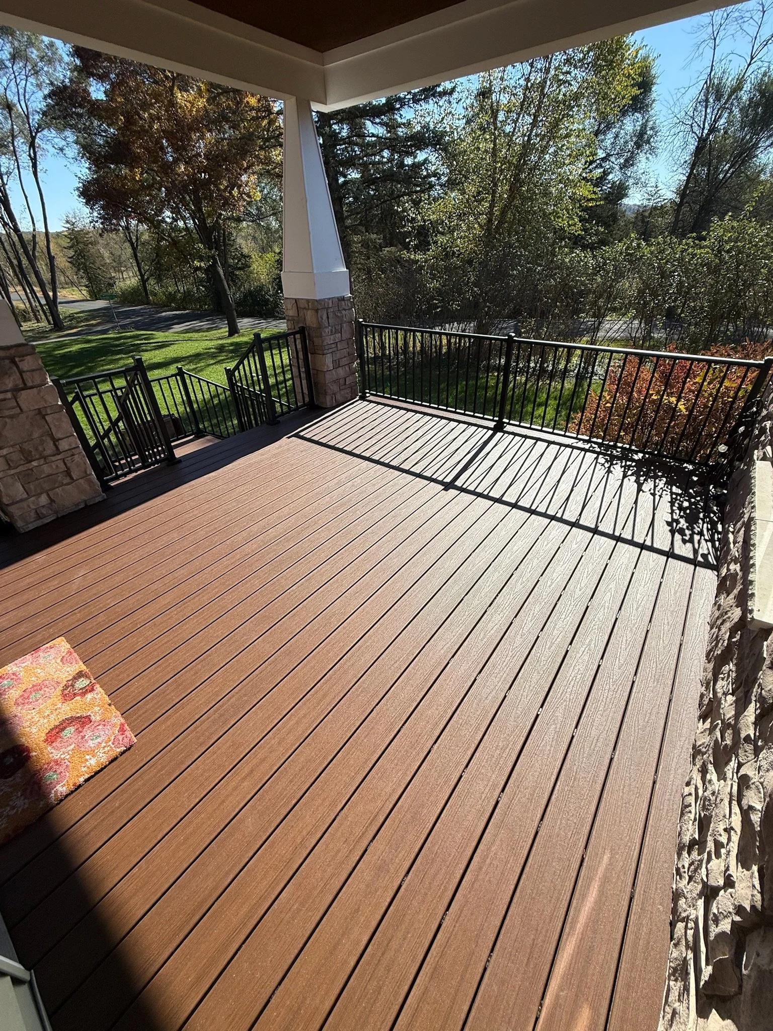 A spacious wooden porch with black metal railing, surrounded by trees and green grass, under a clear blue sky on a sunny day.