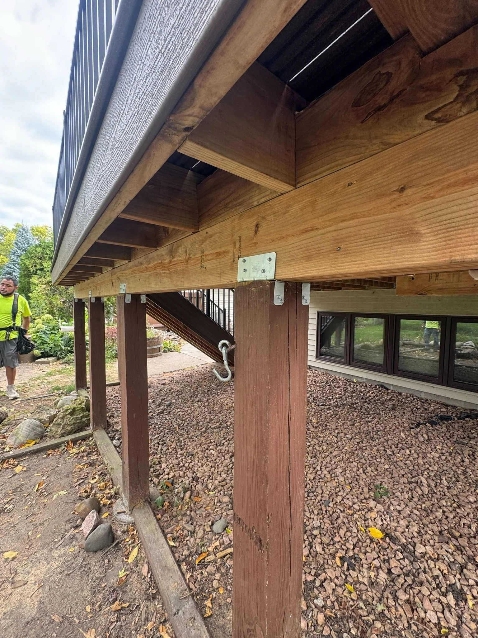 Close-up of a house's elevated wooden deck support structure, showing the underside of the deck with metal brackets and the stairway leading up to it, in a landscaped yard.
