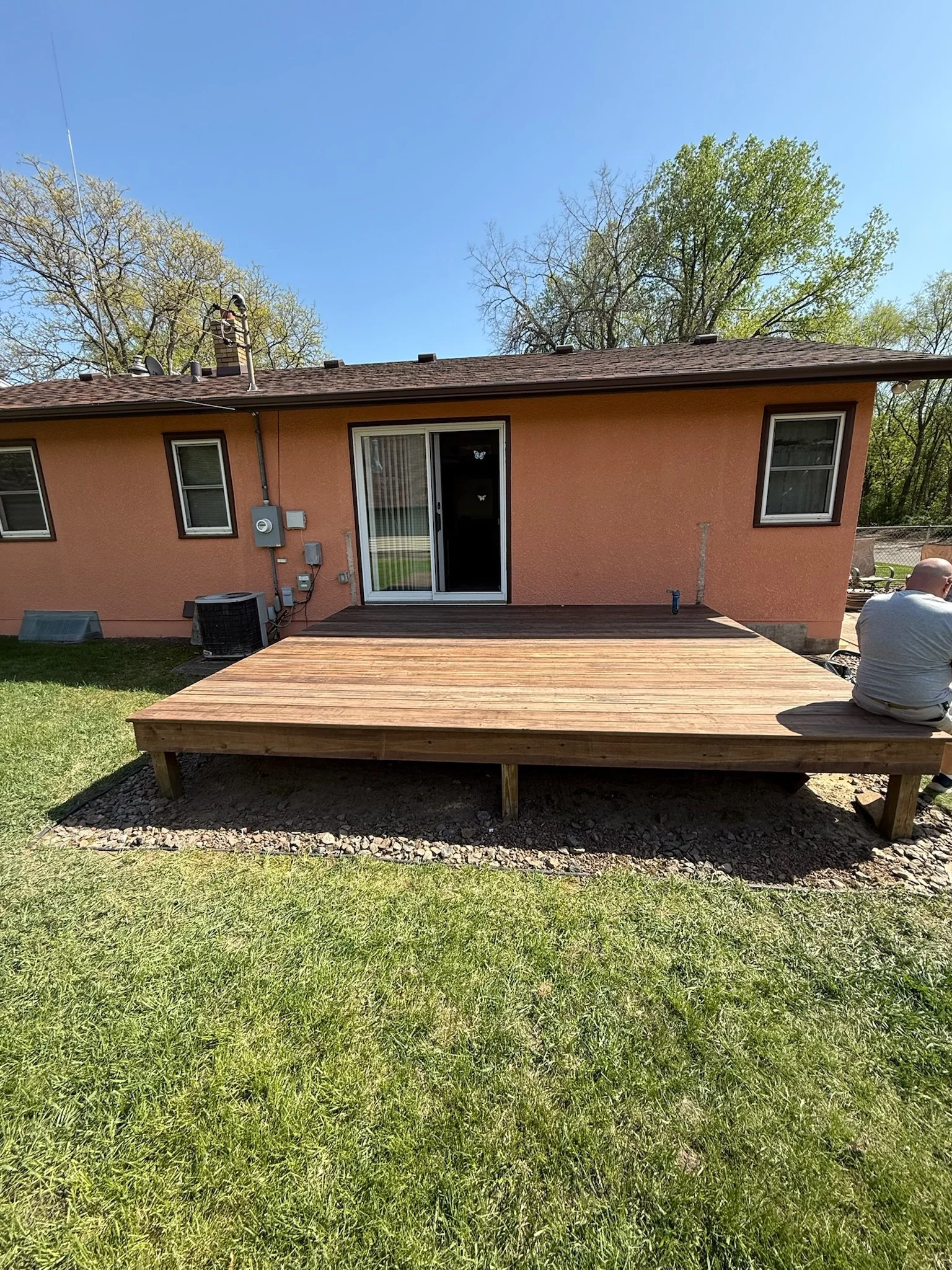 New wooden deck outside a house with a sliding glass door, windows, and a person sitting on the edge of the deck.