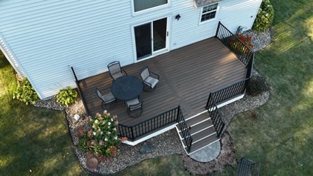 A backyard wooden deck with a round table and four chairs, attached to a white house, surrounded by grass and landscaping.