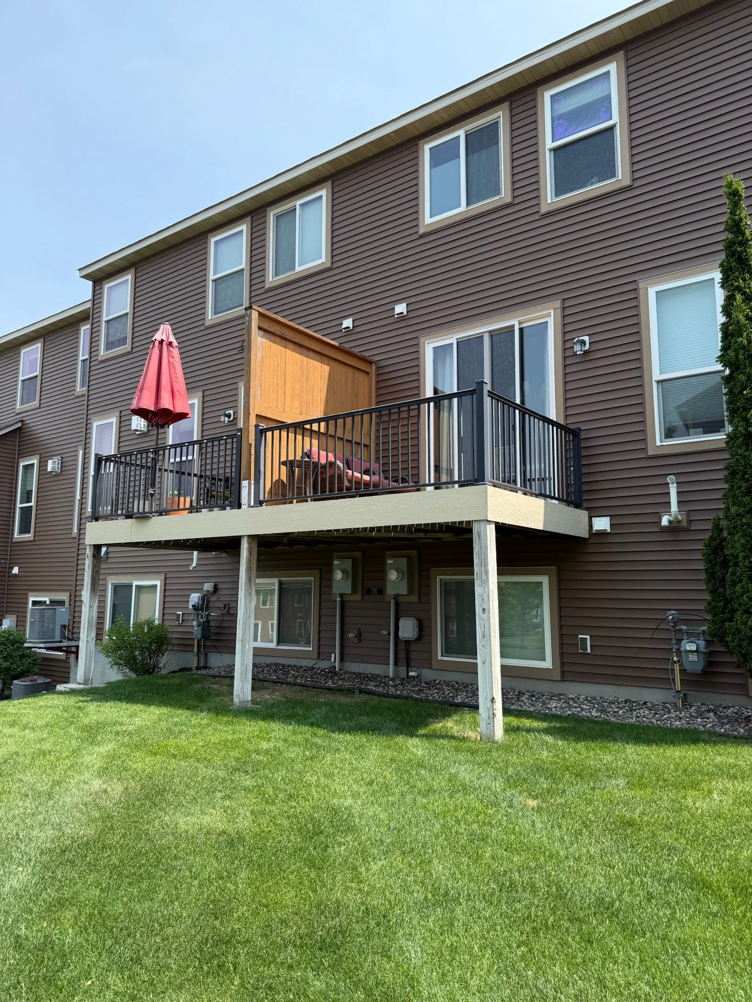 Back of a multi-story residential building with a patio balcony, wooden privacy panel, patio furniture, umbrella, green lawn, and utility meters.