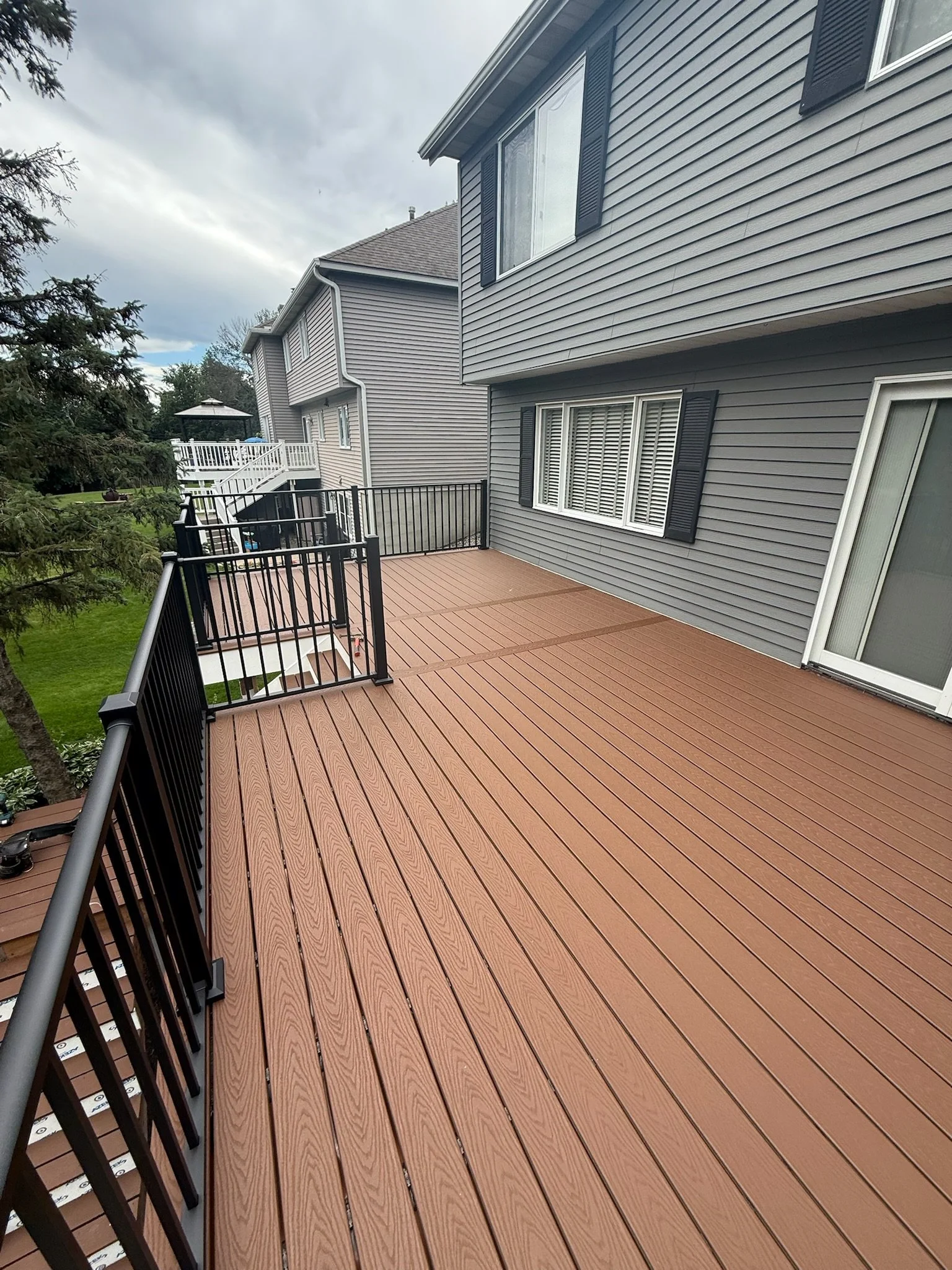 A spacious outdoor deck with brown wooden flooring, black metal railing, attached to a grey house with black shutters and white window frames, overlooking a green yard and neighboring houses under a cloudy sky.