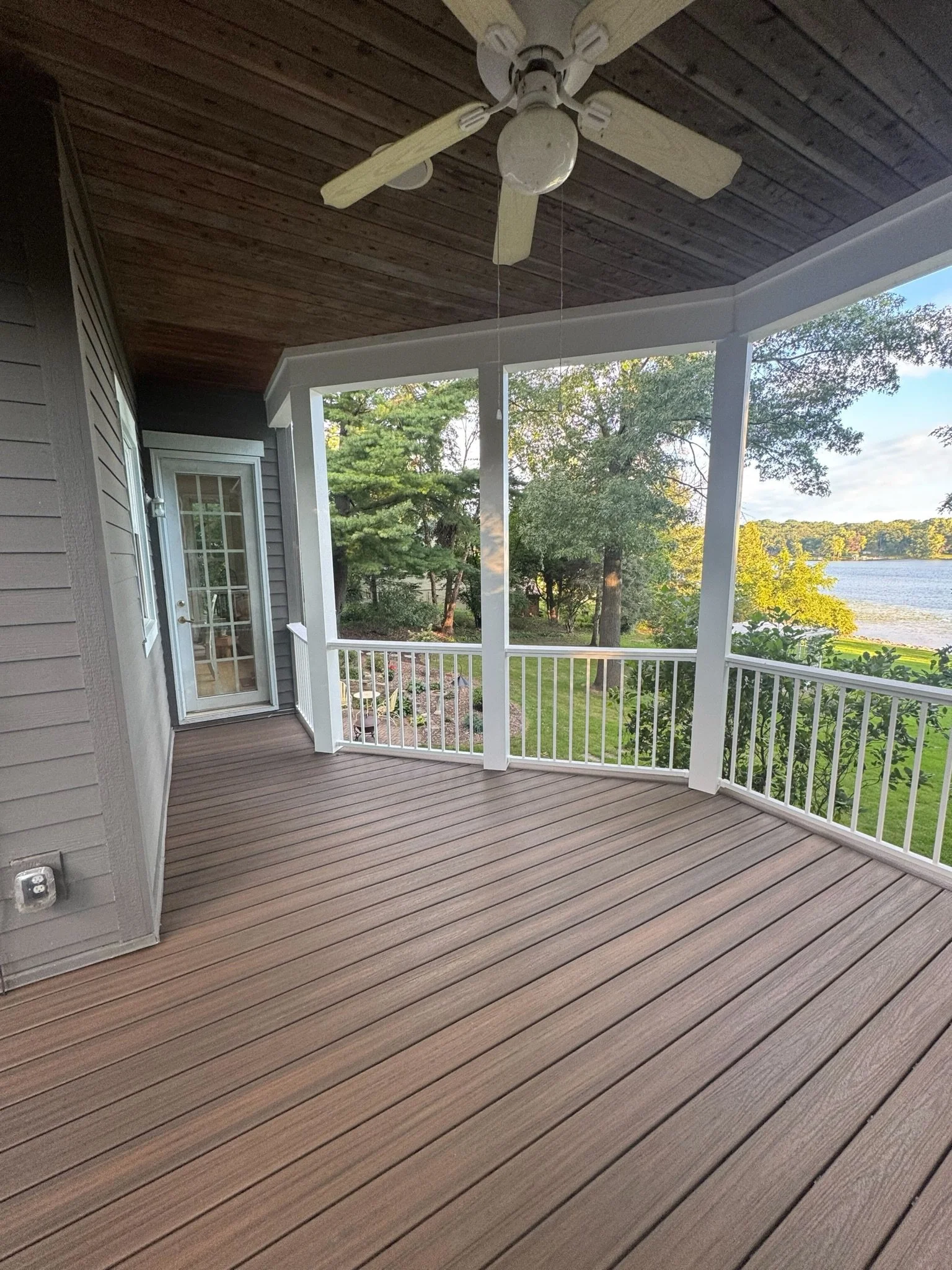 Empty covered porch with wood floor, ceiling fan, and railing overlooking a yard and a lake.