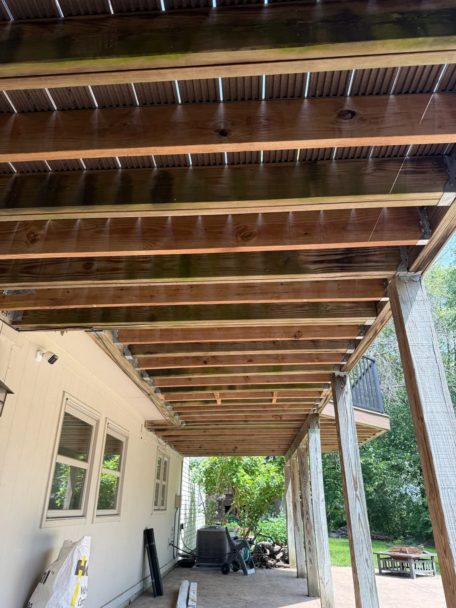 Under construction wooden porch ceiling with exposed beams and support posts, next to a house with multiple windows, outdoor furniture, and trees in the background.