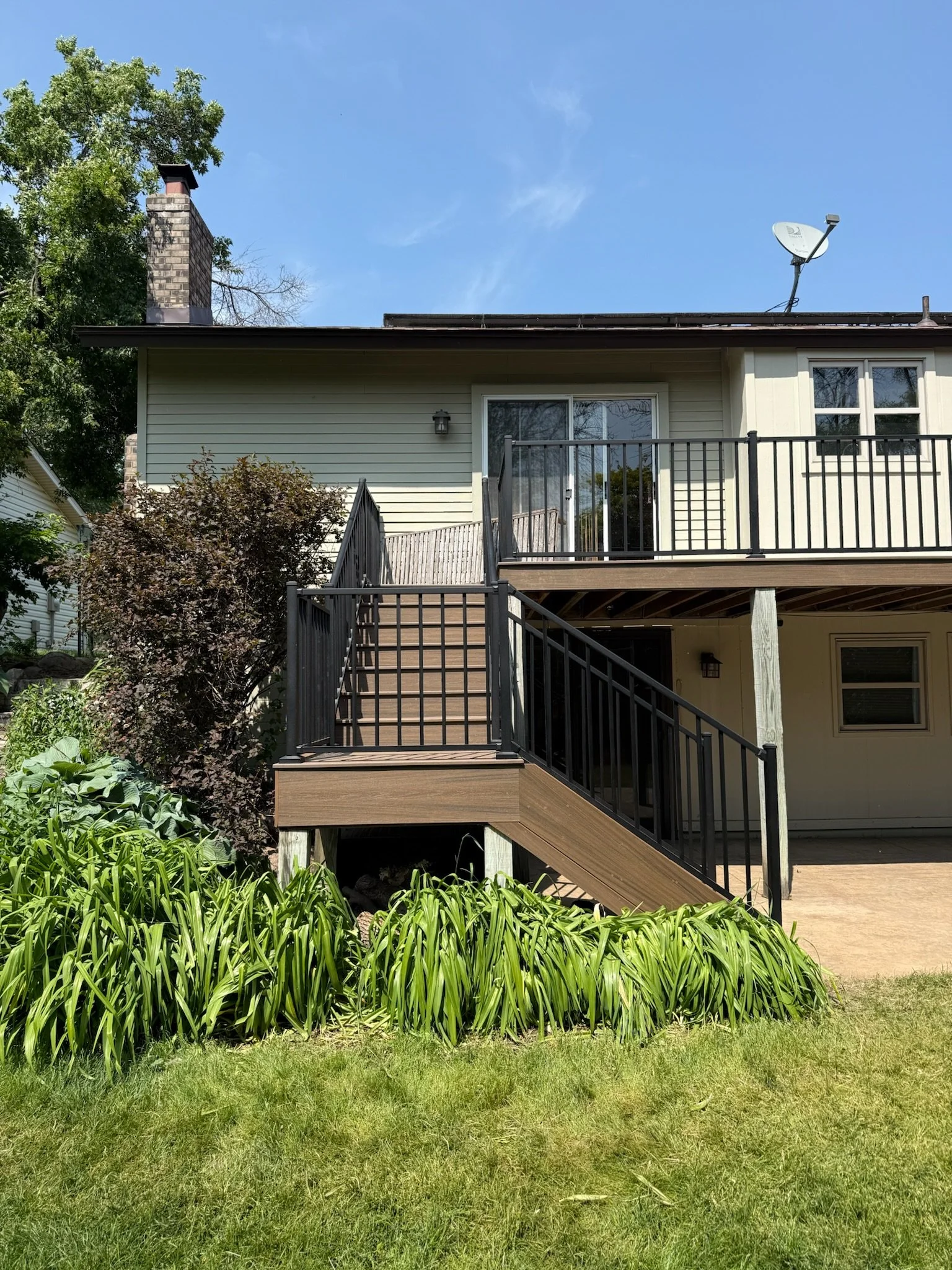 Backyard view of a house with a wooden deck, black railing, and stairs leading down to a grassy yard, surrounded by green plants and trees under a blue sky.