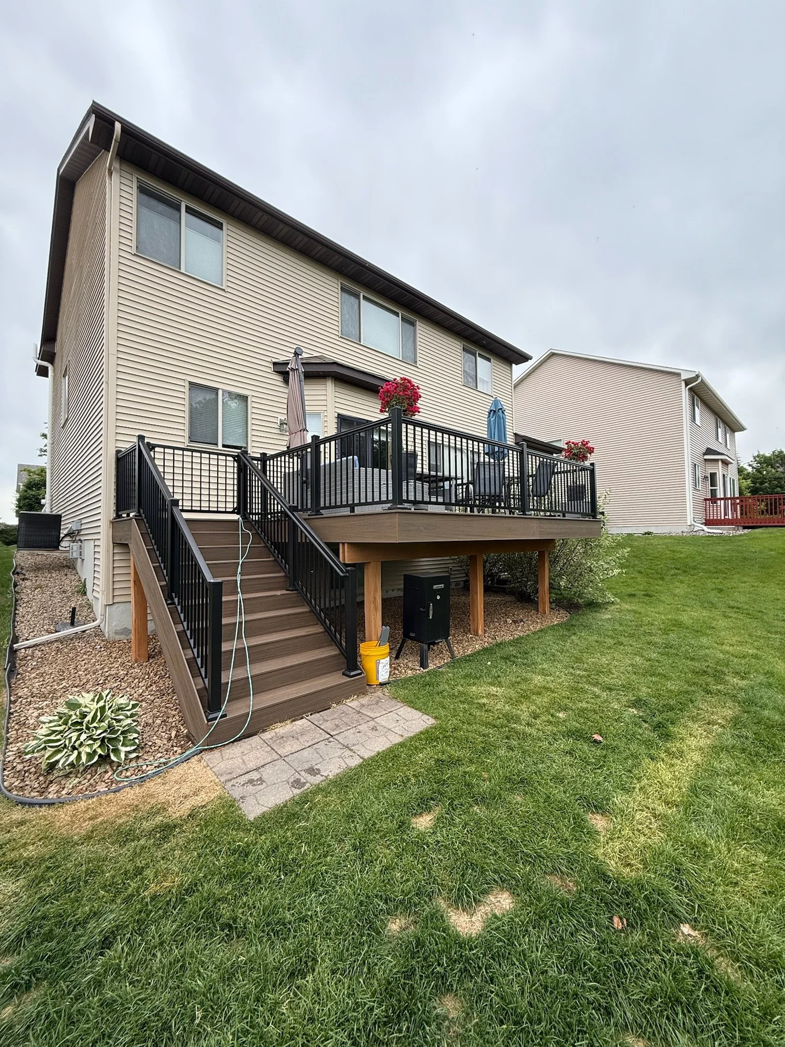 Backyard view of a two-story house with a decorated wooden deck, stairs leading down to a grassy yard, and potted plants with flowers on the deck.