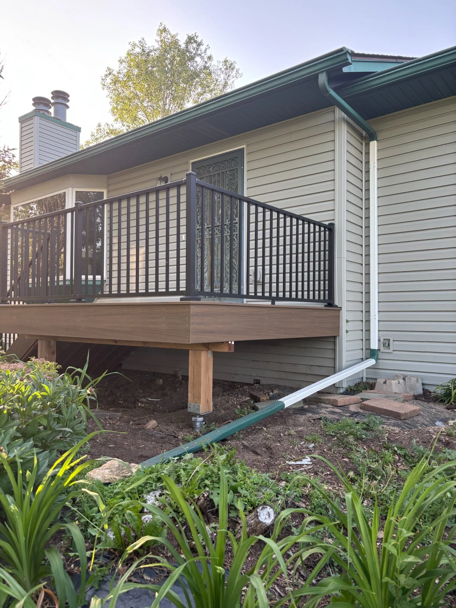 Back porch with newly built wooden deck and black metal railing, located on the second story of a house with beige vinyl siding and green roof trim; part of the drainage system visible on the ground, surrounded by plants and garden area.