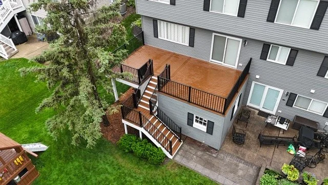 An aerial view of a multi-level deck with wooden flooring and black railings attached to a gray residential building. The deck has stairs leading down to a backyard with a green lawn, trees, and outdoor furniture on a paved patio.
