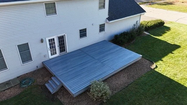 An outdoor metal deck attached to the back of a white house with stairs leading down to a gravel area, surrounded by grass and bushes.
