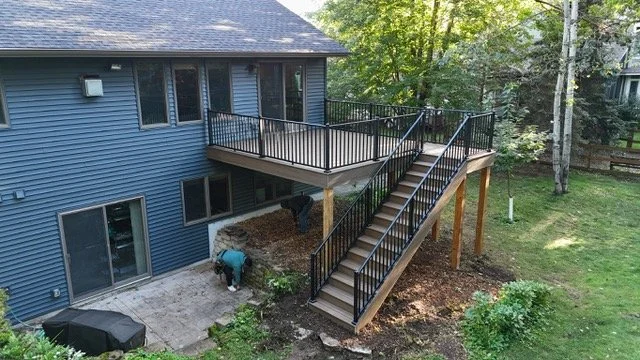 Backyard of blue house with wooden deck, stairs, and black metal railing, surrounded by trees and grass.