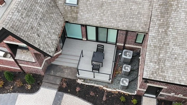 View of a second-floor balcony with outdoor table and chairs, accessed through large sliding glass doors, surrounded by brick building walls, with air conditioning units below and a patterned walkway in the courtyard.