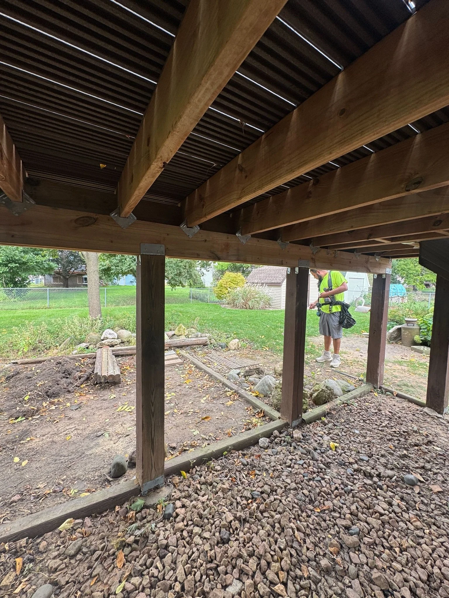 View under a wooden deck with support beams and a person in a yellow safety vest inspecting the area, surrounded by grass, trees, and a small garden.