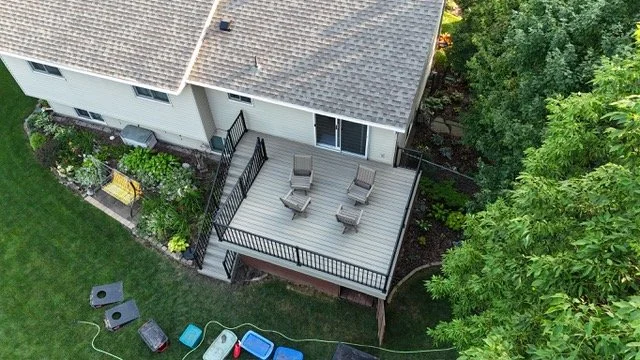 Aerial view of a house's backyard deck with four lounge chairs, surrounded by green trees and lawn, with garden beds and outdoor toys.
