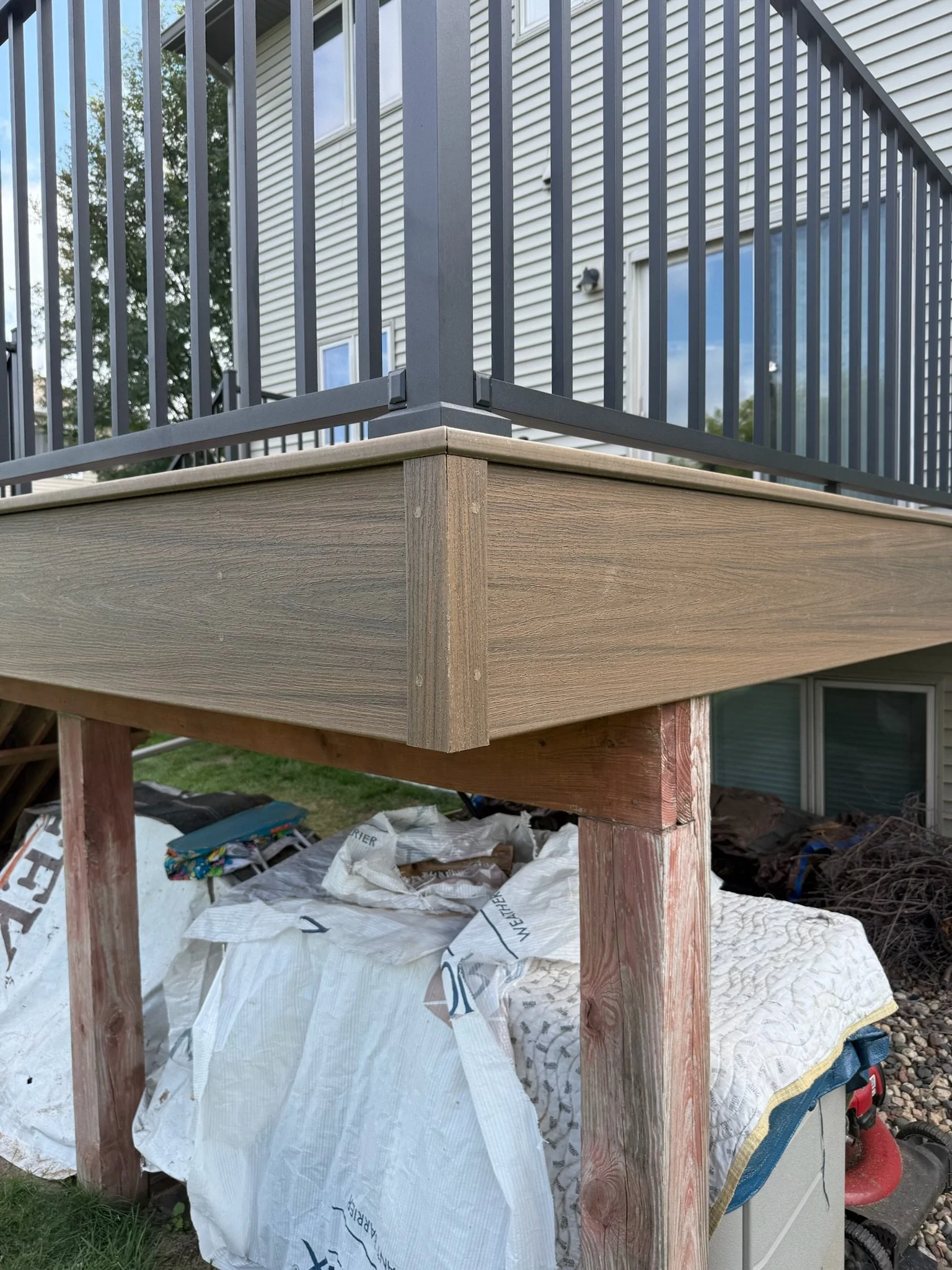 Close-up of a home deck under construction with wooden frame and black metal railing. Construction materials and debris are beneath the deck.