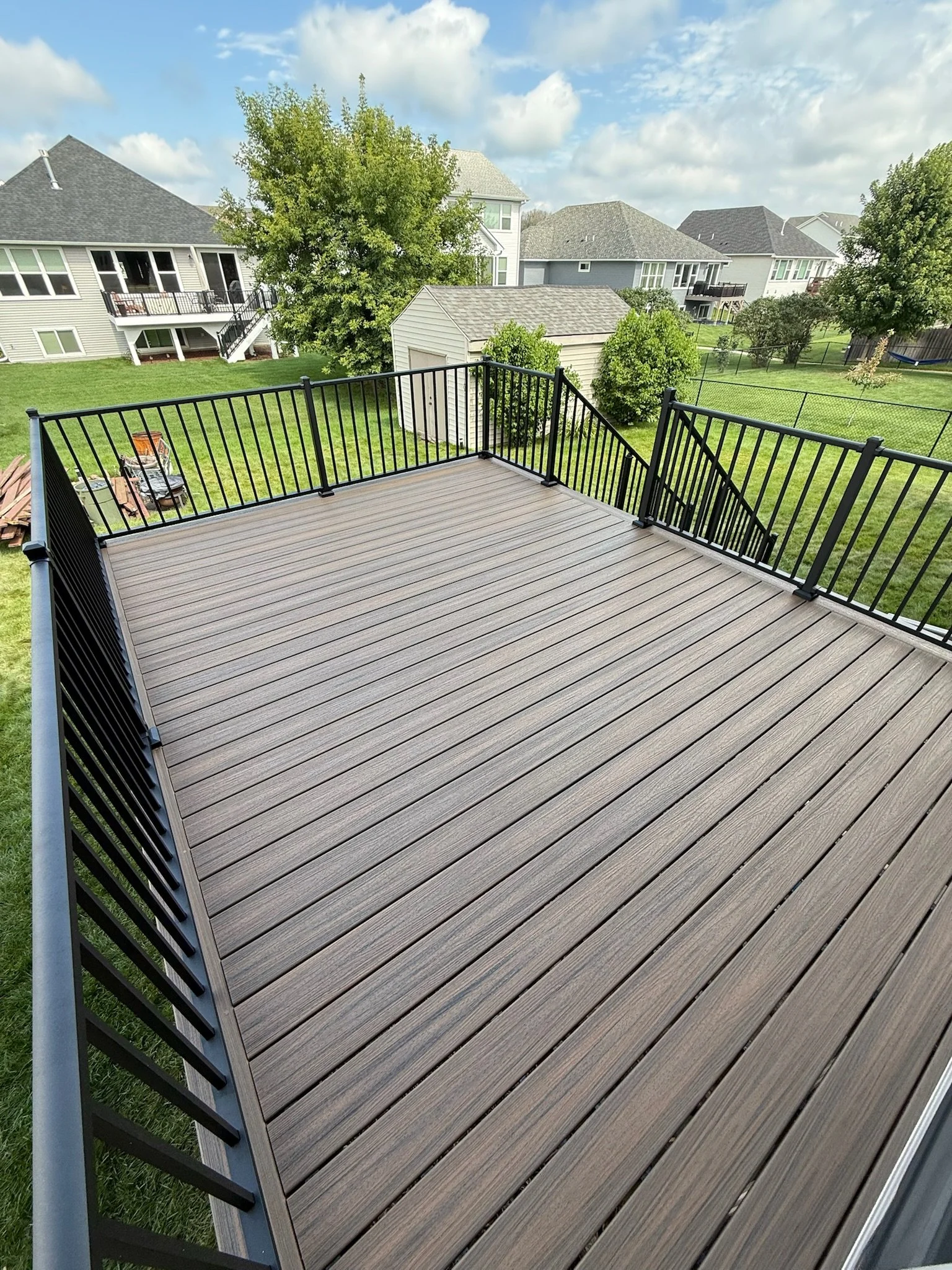 A new wooden deck with metal railing in a backyard with green grass, trees, and neighboring houses under a partly cloudy sky.