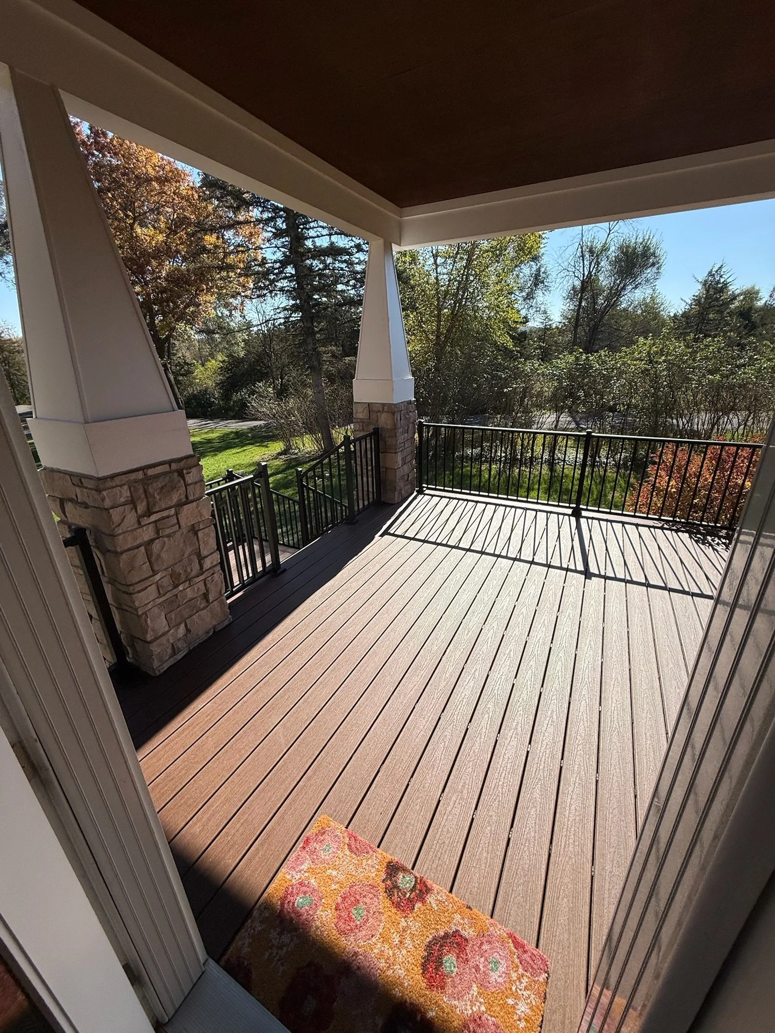 View of a wooden porch or balcony with a black metal railing, stone pillars, and a colorful floral doormat, overlooking green trees and a blue sky.