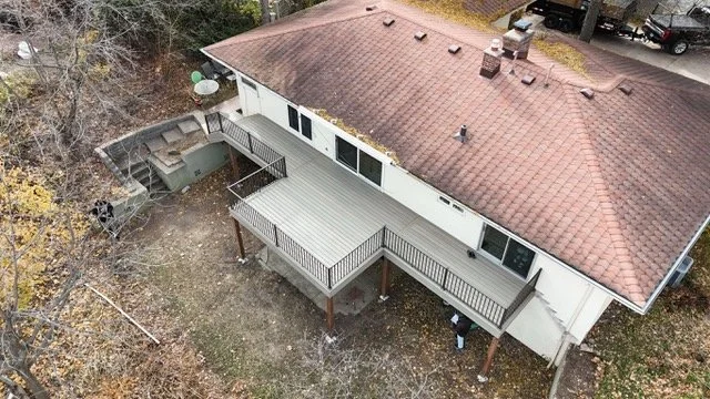 An aerial view of a two-story house with a large roof, upper and lower decks, a backyard with some trees and outdoor equipment, and vehicles parked nearby.