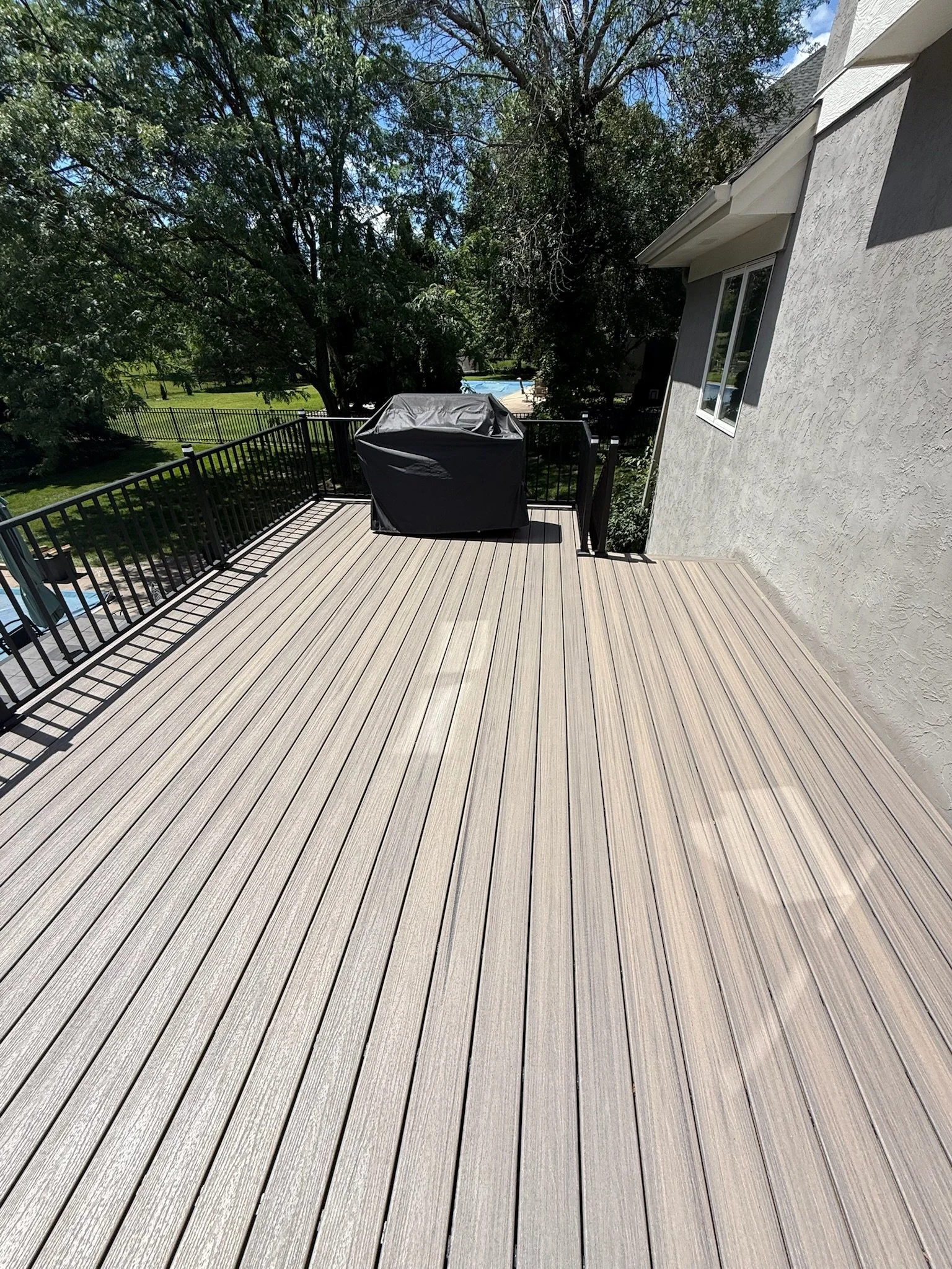 An outdoor wooden deck attached to a beige house with a window, surrounded by trees. There is a black covered barbecue grill on the deck and black metal railing on the left side.