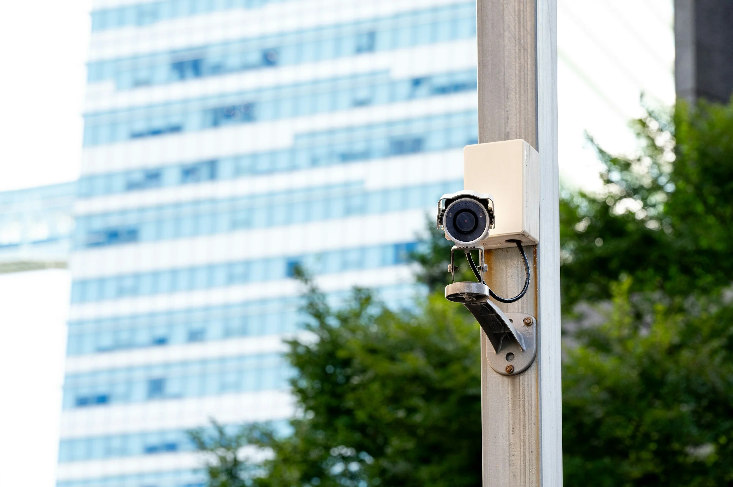 Close-up of an outdoor security camera mounted on a metal pole with a modern glass building and green trees in the background.