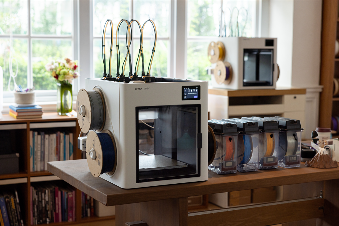 A 3D printer with spools of filament in various colors on a wooden table in a well-lit room.