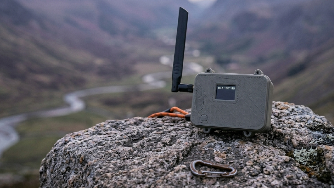A rugged, portable GPS device with an antenna, placed on a large rock in a mountainous landscape with a winding river in the background.