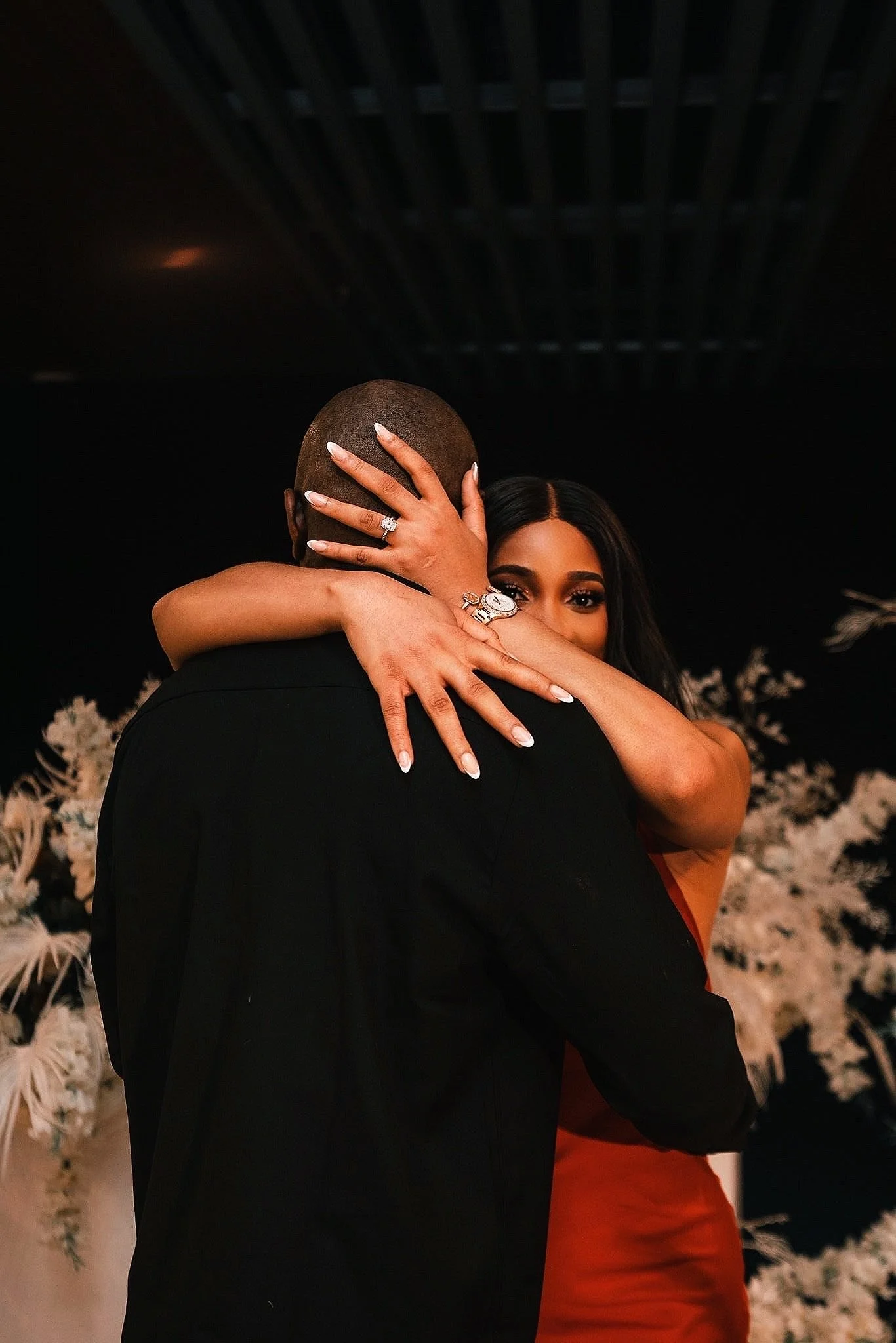A woman and a man hugging each other, with the woman looking at the camera. The woman has long dark hair, is wearing a ring and a watch, and has her arms around the man's head. The man is bald and wearing a black suit, facing away from the camera. The background features a floral arrangement.