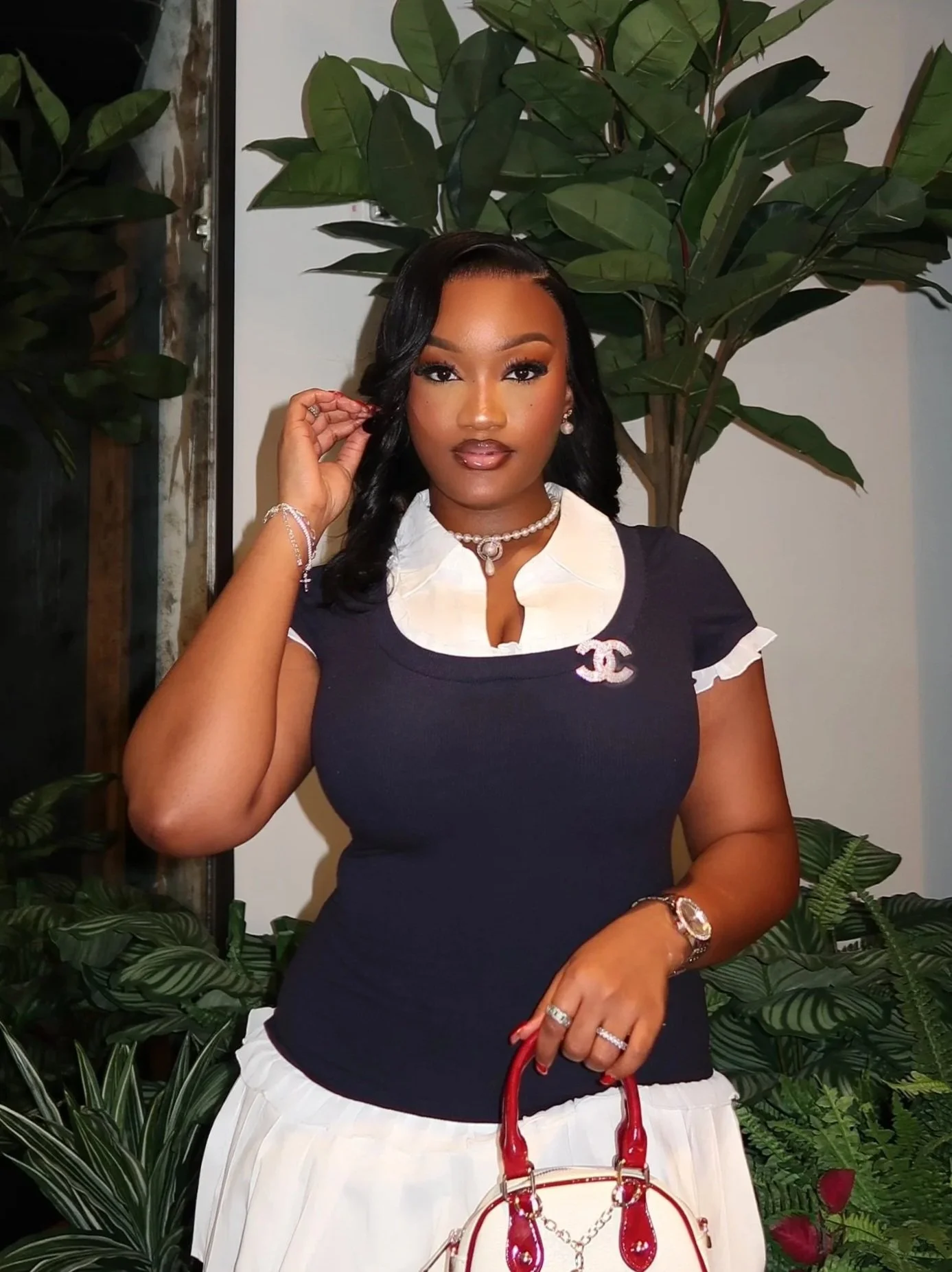 A woman with dark hair and brown skin wearing a black and white top, accessorized with jewelry, holding a white and red handbag, standing in front of green plants.
