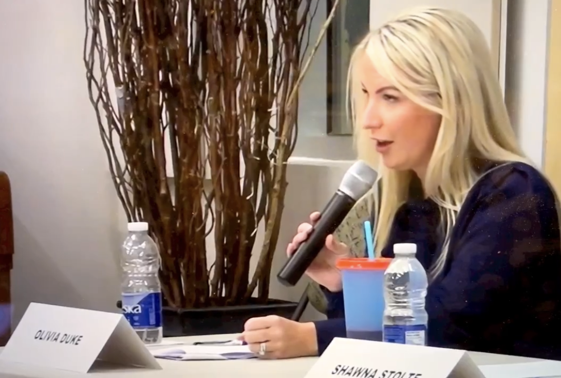 A woman with long blonde hair speaking into a microphone at a panel or meeting table. There are water bottles, a paper cup with a straw, and nameplates with "Olivia Duke" and "Shawnna Stoute" on the table.