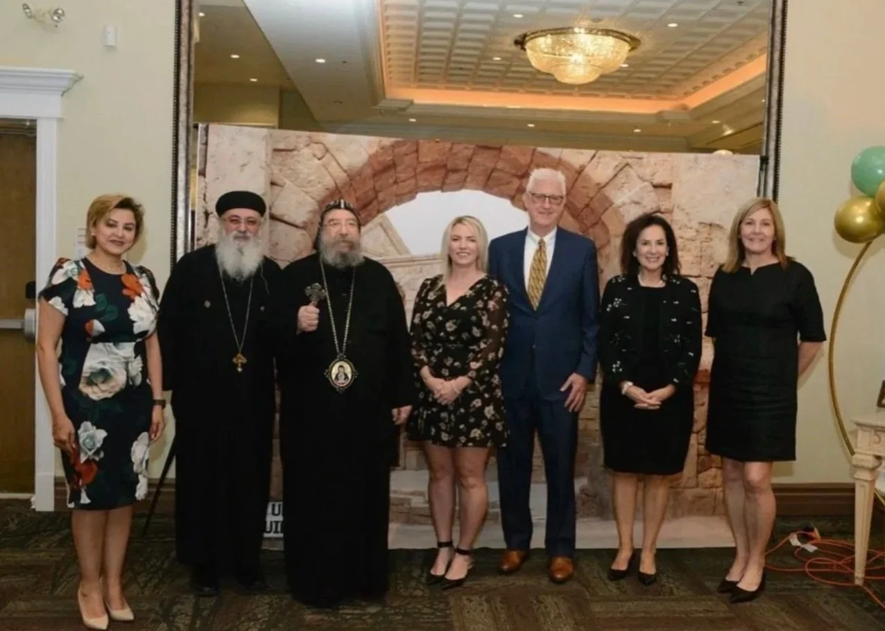 A group of seven people standing in a line in front of a decorative backdrop with an arch and stone texture, inside a banquet hall or event space. They are dressed in formal and semi-formal attire.