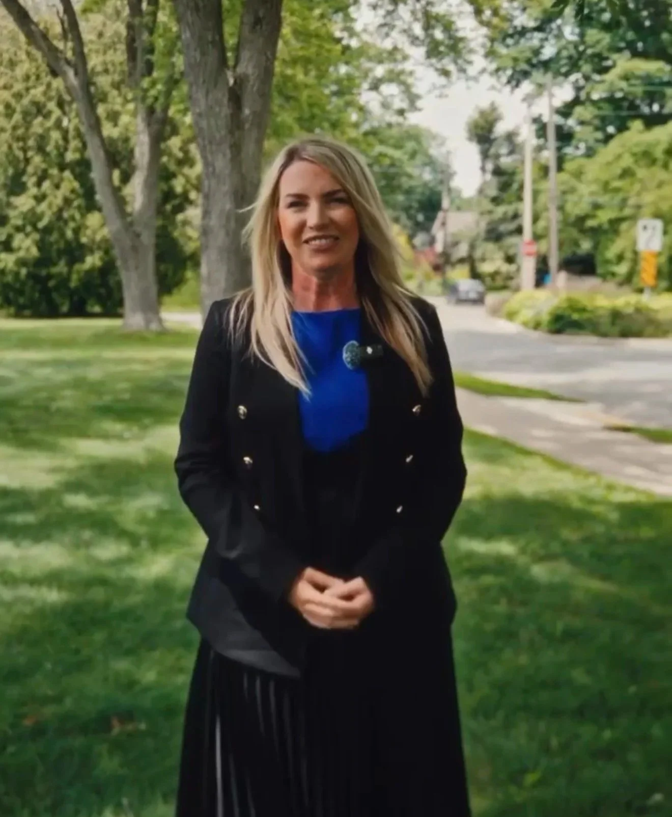 A woman with long blonde hair wearing a blue shirt and a black jacket standing outdoors on grass with trees and a street in the background.